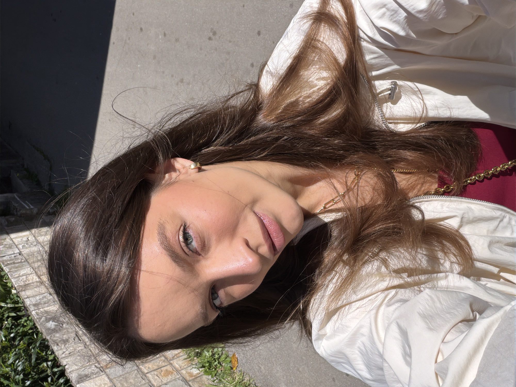 A close-up, high-angle portrait captures a young woman outdoors under bright sunlight. The image is oriented with the subject's head towards the lower right and her body extending diagonally upwards. She has long, dark brown hair that frames her face and spreads around her. Her eyes are blue-green, and she wears a soft, serene smile with slightly parted lips. Her skin shows a natural glow, particularly on her cheeks and nose.
She is dressed in a light-colored (off-white or cream) jacket, which is unzipped, revealing a glimpse of a maroon or dark red top underneath. Gold-toned chain accessories are visible around her neck and near her shoulder, and she wears a small gold earring.
Strong, direct sunlight casts a sharp shadow across the left side of her face and onto the paved ground behind her, suggesting a clear day around midday or early afternoon. The background consists of a grey paved surface, green foliage (bushes or grass) in the corners, and a low stone or brick wall in the bottom left. The overall setting appears to be a casual outdoor location, possibly in Budva, Montenegro, given the context. There is no visible text in the image.