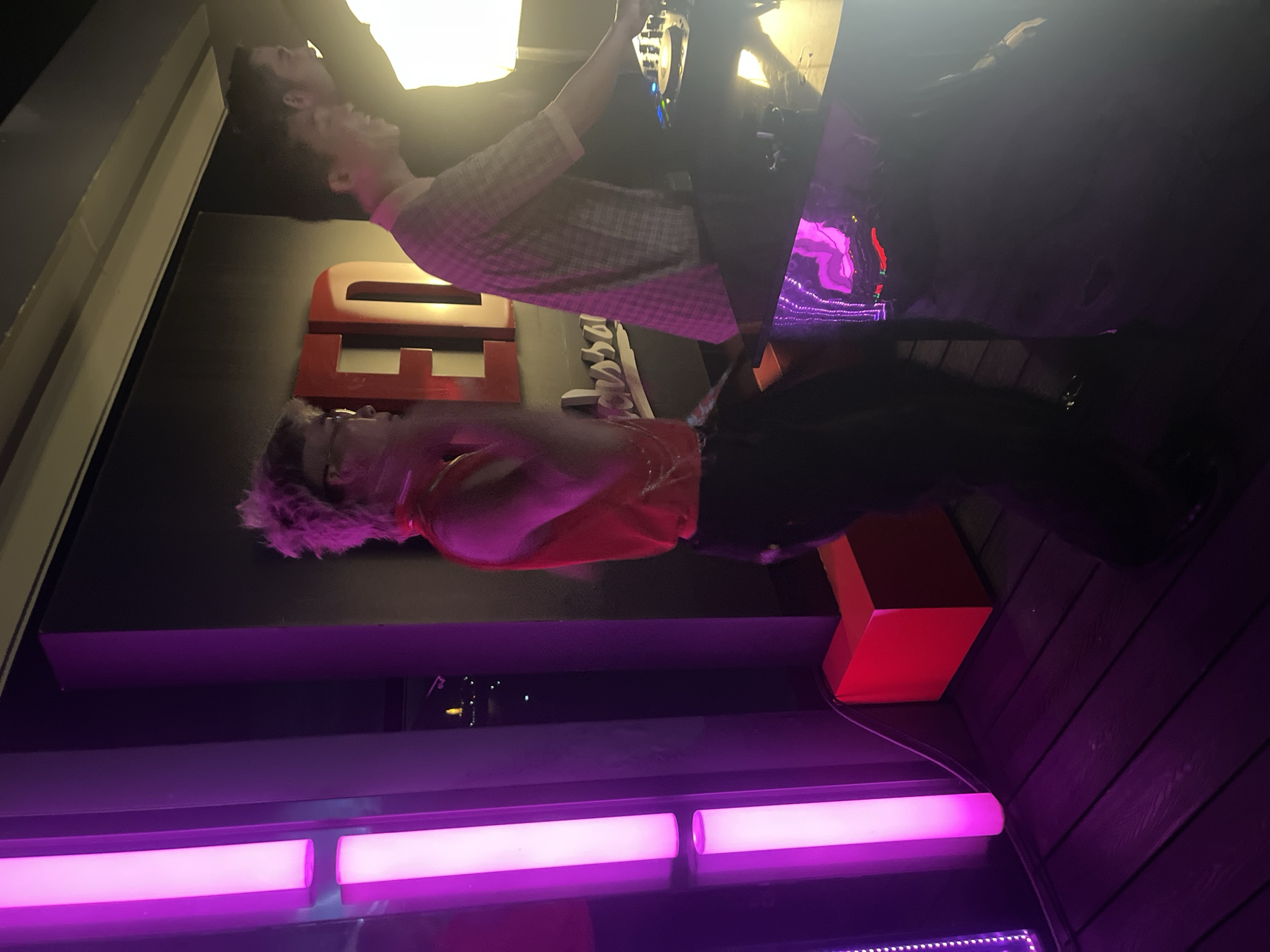 The image captures a vibrant nightlife scene at "RED" nightclub, located on Võ Nguyên Giáp Street in Đà Nẵng, Vietnam. The foreground is dominated by tall, vertical purple LED lights, casting a colorful glow on the dark wooden floor. A person with spiky blonde hair, wearing a bright red sleeveless top and dark pants, is in the center, seemingly dancing or gesturing. To the right, another individual, wearing a checkered shirt, appears to be operating DJ equipment. Behind them, the distinctive "RED" logo is visible on the wall, illuminated in red. The overall ambiance suggests a lively party or club event, with the purple and red lighting contributing to a dynamic and energetic atmosphere. The reflection on the DJ booth shows some abstract purple and pink shapes, adding to the visual interest of the scene.