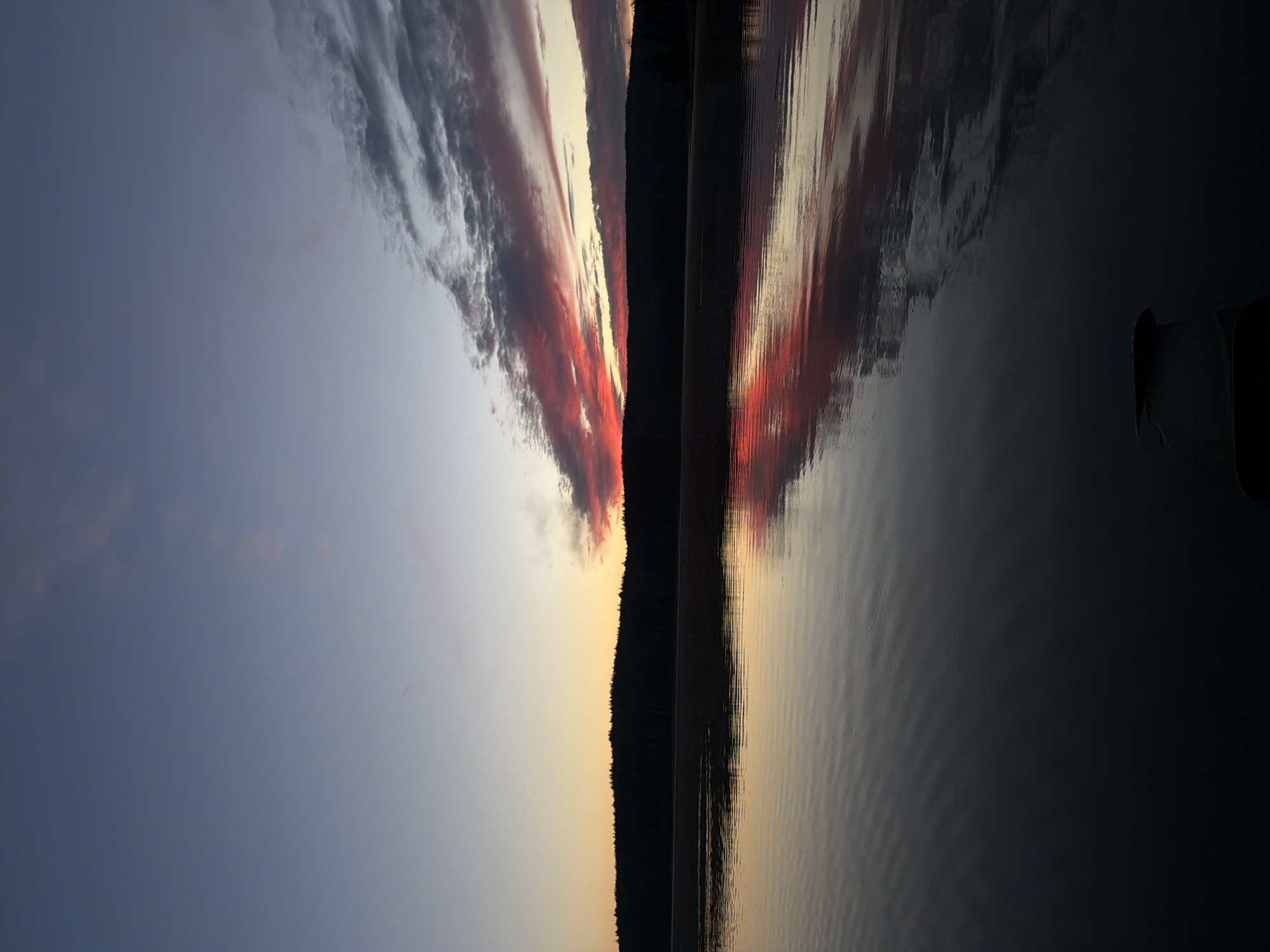 A vertically oriented photograph captures a sunset reflected on water. The sky transitions from a deep blue on the left to an orange hue near the center, where a line of silhouetted trees marks the horizon. Red and grey clouds are present above the trees. The right side of the image shows the water's surface, reflecting the colors of the sky and clouds. The water has small ripples on the surface. A portion of a boat is visible in the lower right corner.
While the request mentioned Chaikovsky, Russia, the photo alone does not confirm that location.