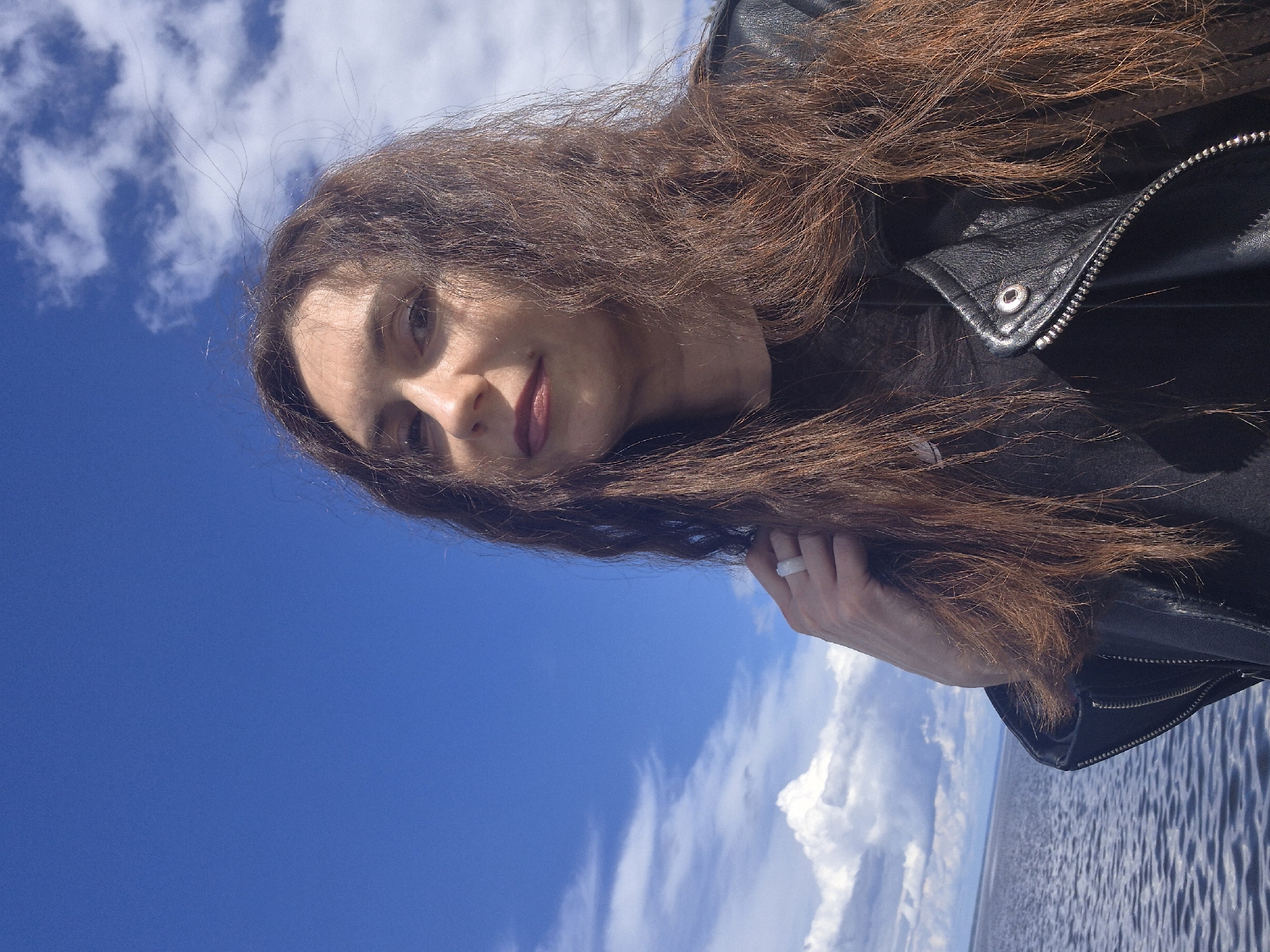 A woman is seen in a medium shot against a backdrop of a bright blue sky with scattered white clouds and a glimpse of what appears to be a body of water. She has long, wavy brown hair and is wearing a black leather jacket. The jacket's metallic zipper and a button are visible. Her face is turned towards the camera with a subtle expression. She is wearing a ring on her hand which is also visible. The location is Unknown, Unknown.