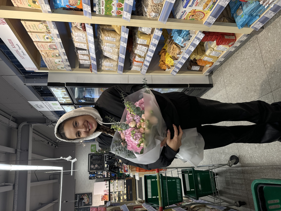 The image depicts a female individual standing in an aisle of a grocery store in Odintsovo, Russia. She is smiling, looking directly forward, and wears a black jacket over a white hooded sweatshirt with the hood up, paired with black trousers. She holds a large bouquet of pink hydrangeas, small roses, and green foliage, wrapped in translucent white and clear material, with both hands at waist level. To her immediate left, a metal shopping trolley containing two green plastic baskets is visible. To her right, a tall retail shelving unit, constructed with light-colored wood and metal, displays numerous packaged dry goods such as grains and cereals, with Russian labels including "КРУПЫ. КАШИ" (Grains. Porridges) and "ВИТЬБА". Price tags are affixed to the shelves below products. In the background on the left, other store features include a refrigerated display case, a self-checkout area marked by an "АВТОКАССА" sign, and a bakery section displaying cakes and pastries under a "ТОРТЫ, ПИРОЖНЫЕ" sign. The store has a speckled grey tiled floor, and an exposed industrial-style ceiling with overhead fluorescent lighting fixtures.