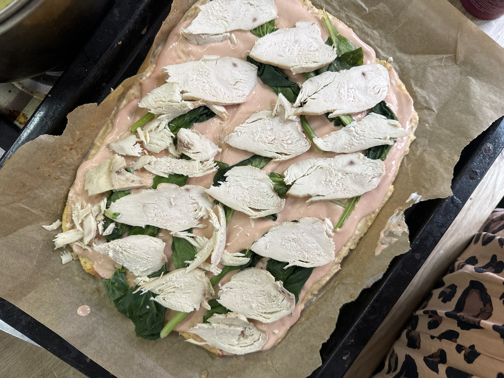 The image shows an uncooked pizza on a baking sheet lined with parchment paper. The pizza appears to have a pink sauce base, topped with spinach and sliced, cooked chicken. The image includes a glimpse of a metal bowl in the top left corner, a black baking sheet edge, and a portion of clothing with a leopard print pattern on the right side. The location cannot be determined from the image. The image does not show anyone.