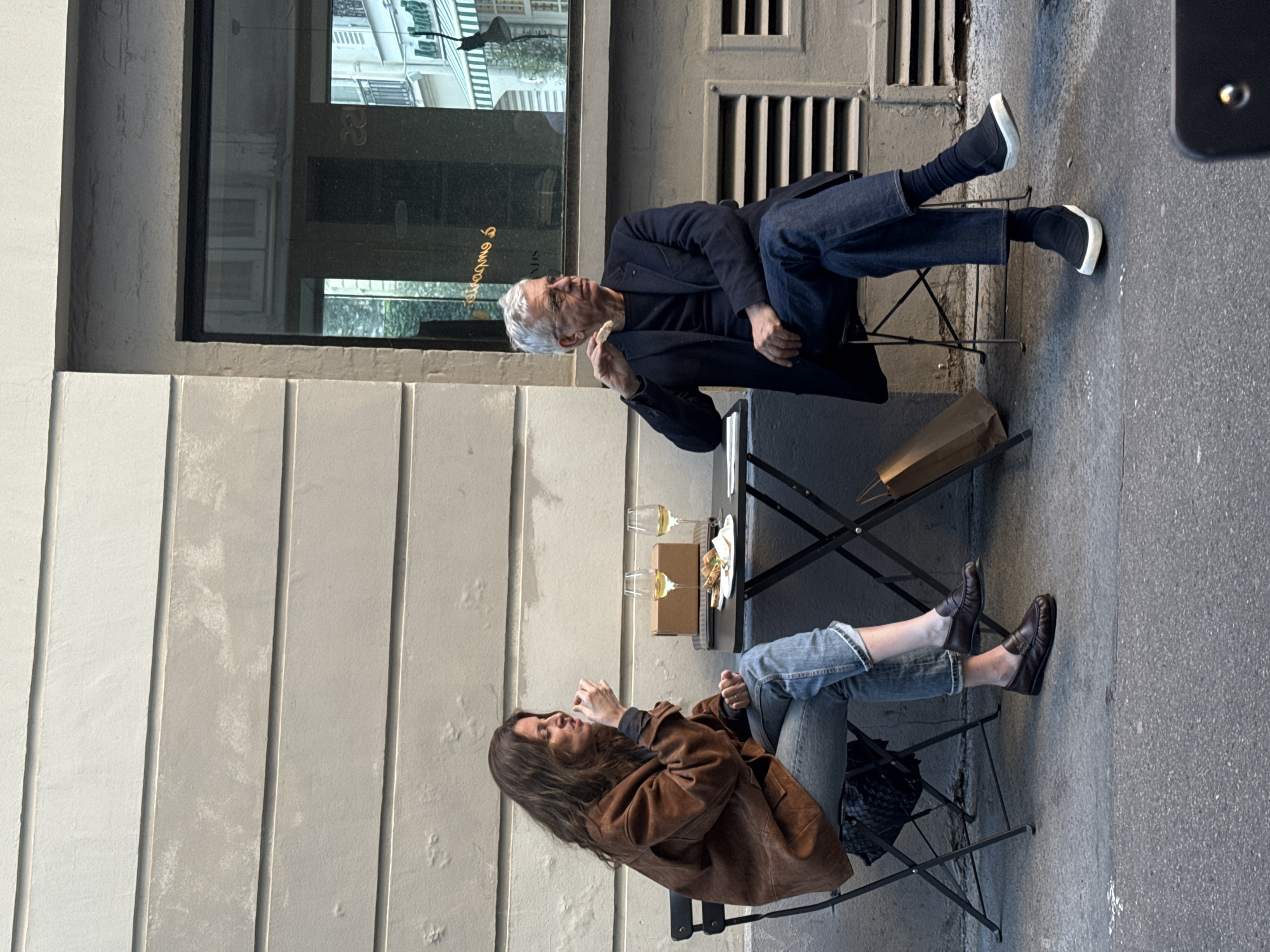 The image depicts two people seated at a small table outside what appears to be a Parisian bistro or cafe. An older man with grey hair and glasses is wearing a dark jacket and jeans, eating something. A younger woman with long, brown hair is wearing a brown jacket and jeans, talking with gestures. On the table are two glasses of white wine and a small plate of food. The background shows a building facade with a window reflecting a Parisian street scene. The setting is a narrow sidewalk next to a building with vertical panels. The scene suggests a casual outdoor meal or conversation in an urban environment.