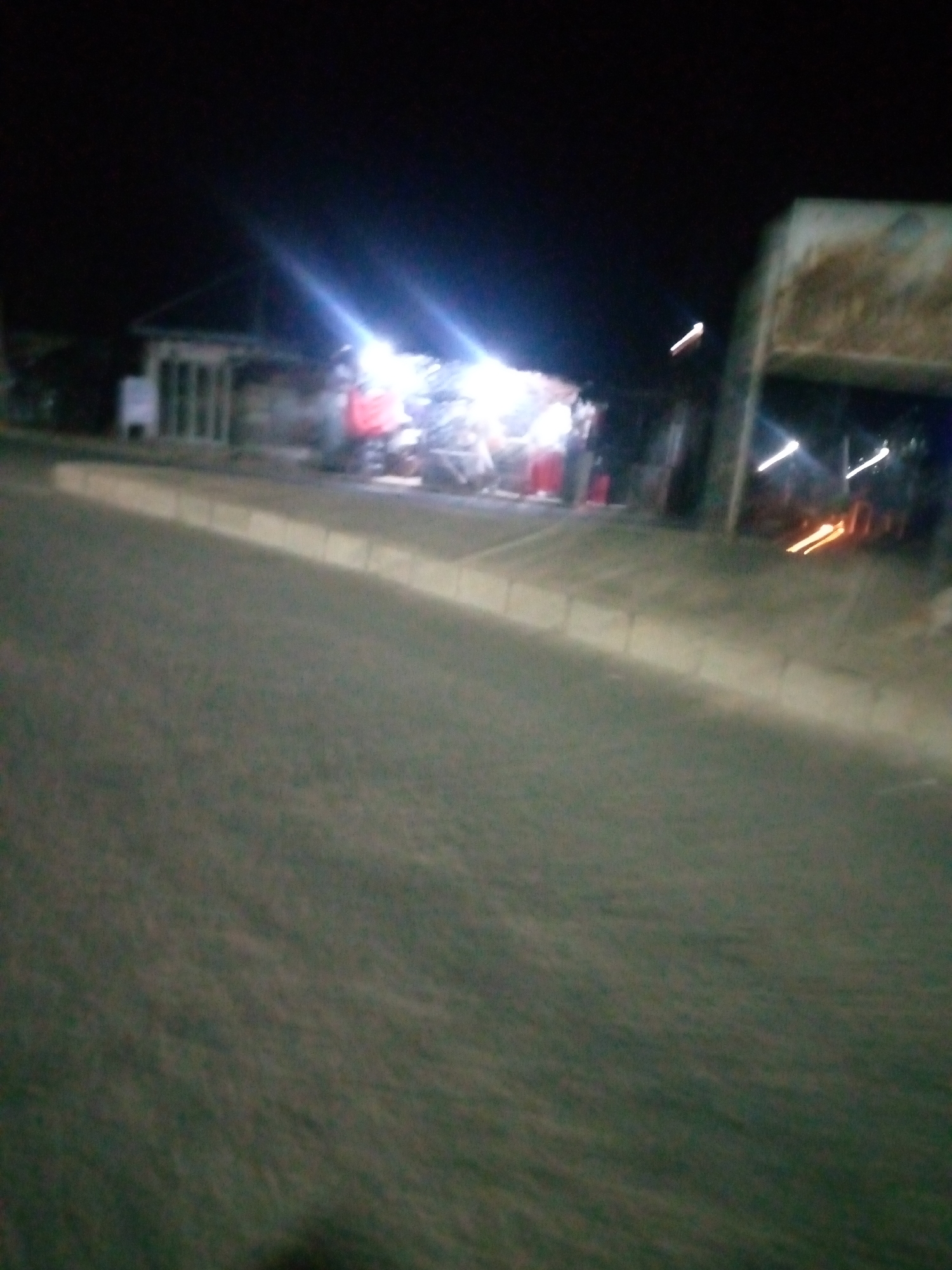 This is a nighttime, blurry, low-light photograph taken from a moving vehicle, likely on a street in Jalingo, Nigeria. The scene depicts a roadside market or gathering area with several stalls illuminated by bright lights, creating significant glare and lens flare. In the background, a building with a sloped roof is visible. The foreground is dominated by the dark, asphalt surface of a road and a light-colored concrete curb. There are some indistinct human figures and objects, possibly motorcycles or carts, clustered around the lit stalls. The overall impression is one of a bustling nocturnal scene at a roadside commercial hub.