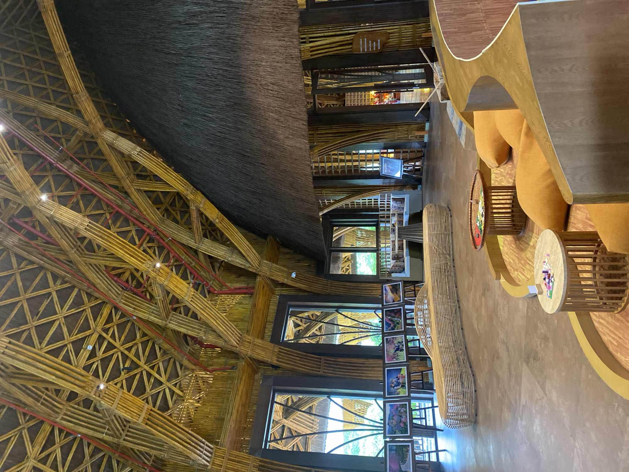 This image captures the interior of a large architectural space located in Púng Luông, Vietnam, with no individuals present. The dominant architectural feature is an elaborate ceiling and structural framework composed extensively of light-toned bamboo poles. The ceiling forms a complex, interwoven lattice of bamboo arranged in triangular patterns, supported by prominent arched bamboo ribs. Red-colored pipes, likely for utility systems, are visible running along portions of the bamboo structure. A substantial section of the roof on the right is covered with dark, shaggy thatch.
Structural columns and wall accents throughout the interior are also constructed from vertical bamboo poles. On the left side, several tall, dark-framed rectangular windows provide natural light and views of green foliage outside; some windows incorporate decorative bamboo patterns within their panes. The floor is a smooth, light brown or beige surface, consistent with polished concrete or large tiles.
In the foreground, a curved, built-in seating area features a wooden base topped with several plump, orange cushions. Two small, circular wooden tables with slatted bases are positioned in front of this seating; one table holds a collection of small, multi-colored game pieces. Further back, a long, undulating bench crafted from light-colored woven rattan or bamboo slats extends across the mid-ground. Along the wall beneath the windows, seven framed artworks, likely photographs depicting landscapes, flowers, and people, are displayed on individual wooden easels. In the background on the right, additional seating with patterned cushions and a round table are partially visible. A rustic wooden sign, bearing text that appears to include "LOBBY," "LOUNGE," and "UNDERGROUND," is affixed to a pillar near an entryway. Recessed lighting fixtures are integrated into the bamboo ceiling structure.