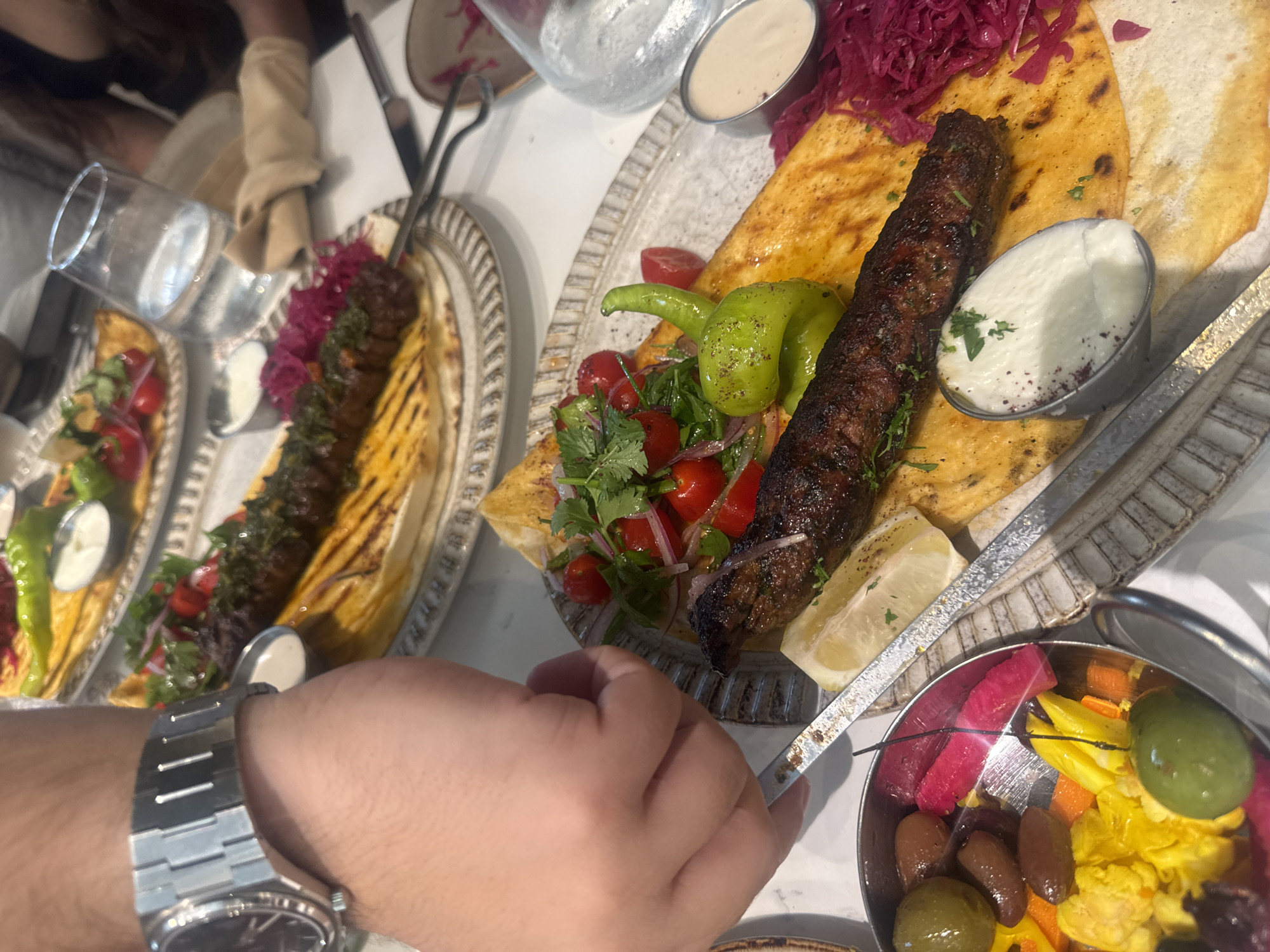 An overhead, medium shot captures a person's left wrist, adorned with a silver watch, as their hand reaches for a meal at a restaurant in Aventura, Florida. The meal is presented on a white plate with a patterned rim, featuring a long, grilled kebob, flatbread, a side of yogurt dip, chopped tomatoes and red onions, a pickled green pepper, and shredded purple cabbage. Another plate with similar food is visible in the background, as is a glass of water and a napkin. The setting appears to be a well-lit indoor dining area.