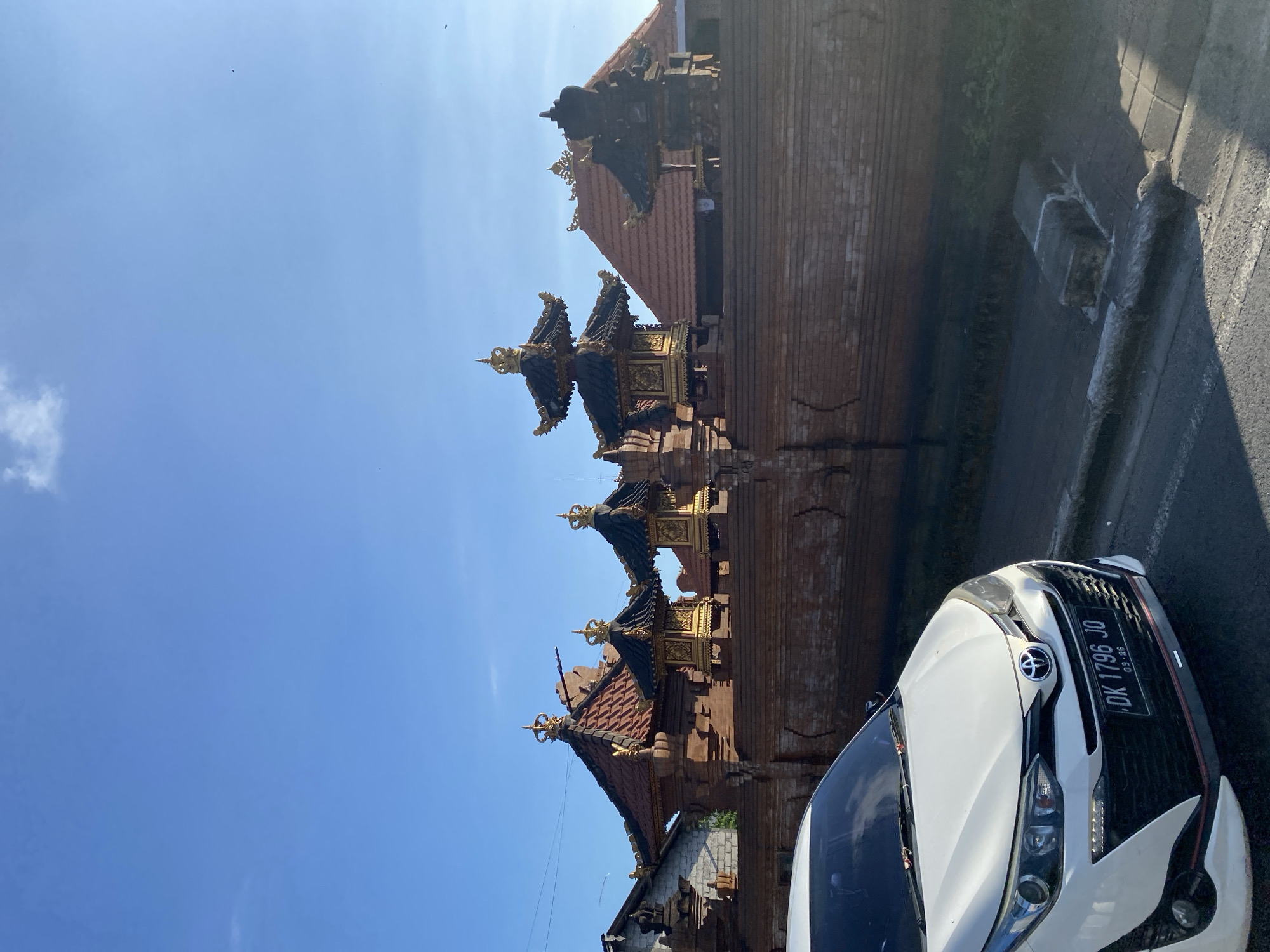 The image shows a brick structure with tiered decorative roofs, likely a traditional Balinese building. A white car is parked in the foreground, partially obscuring the view. The license plate is visible and reads "DK 1796 JO." The sky is clear and blue. The location is North Kuta, Indonesia.