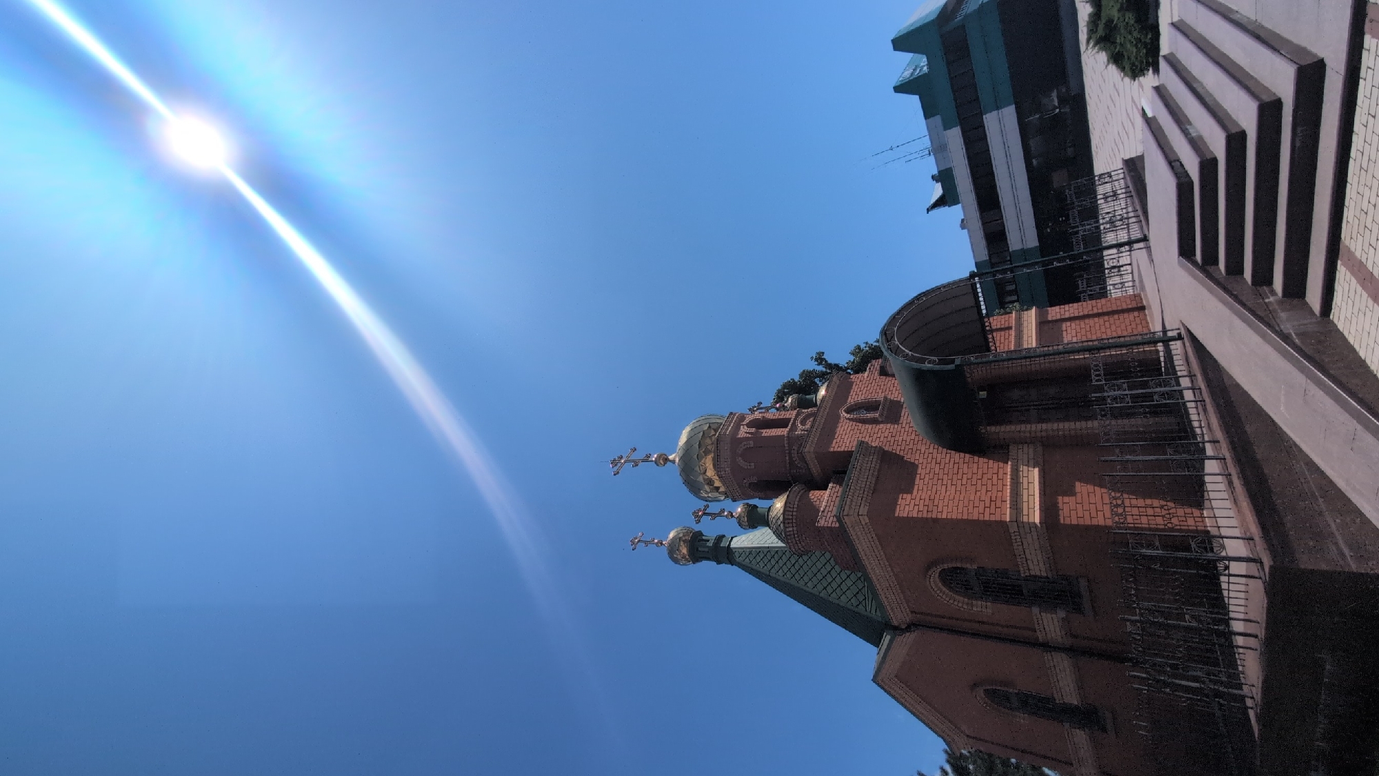 In Izmail, Ukraine, under a bright, sunny sky with visible lens flare, a traditional brick church with a green roof and golden domes stands prominently. The church features ornate golden crosses atop its domes. In front of the church, a paved walkway leads up a set of stone steps, bordered by a decorative black metal fence. To the right of the church, a modern building with a distinctive green and dark-colored facade is visible. The scene is devoid of people.