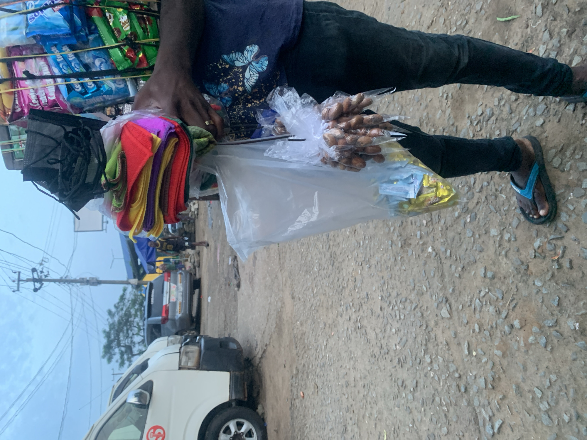 An individual stands in a gravelly urban environment, likely a marketplace in Elelenwo, Nigeria, carrying a large assortment of goods. The person is wearing dark pants and blue flip-flops. They are holding a large bundle of colorful cloths in plastic wrap, along with packages of snacks and face masks. To the left of the individual, two white vans are parked, and a silver SUV is visible in the background. The sky is overcast, suggesting either early morning or late evening. The scene depicts a street vendor engaged in their trade amidst everyday urban activity.