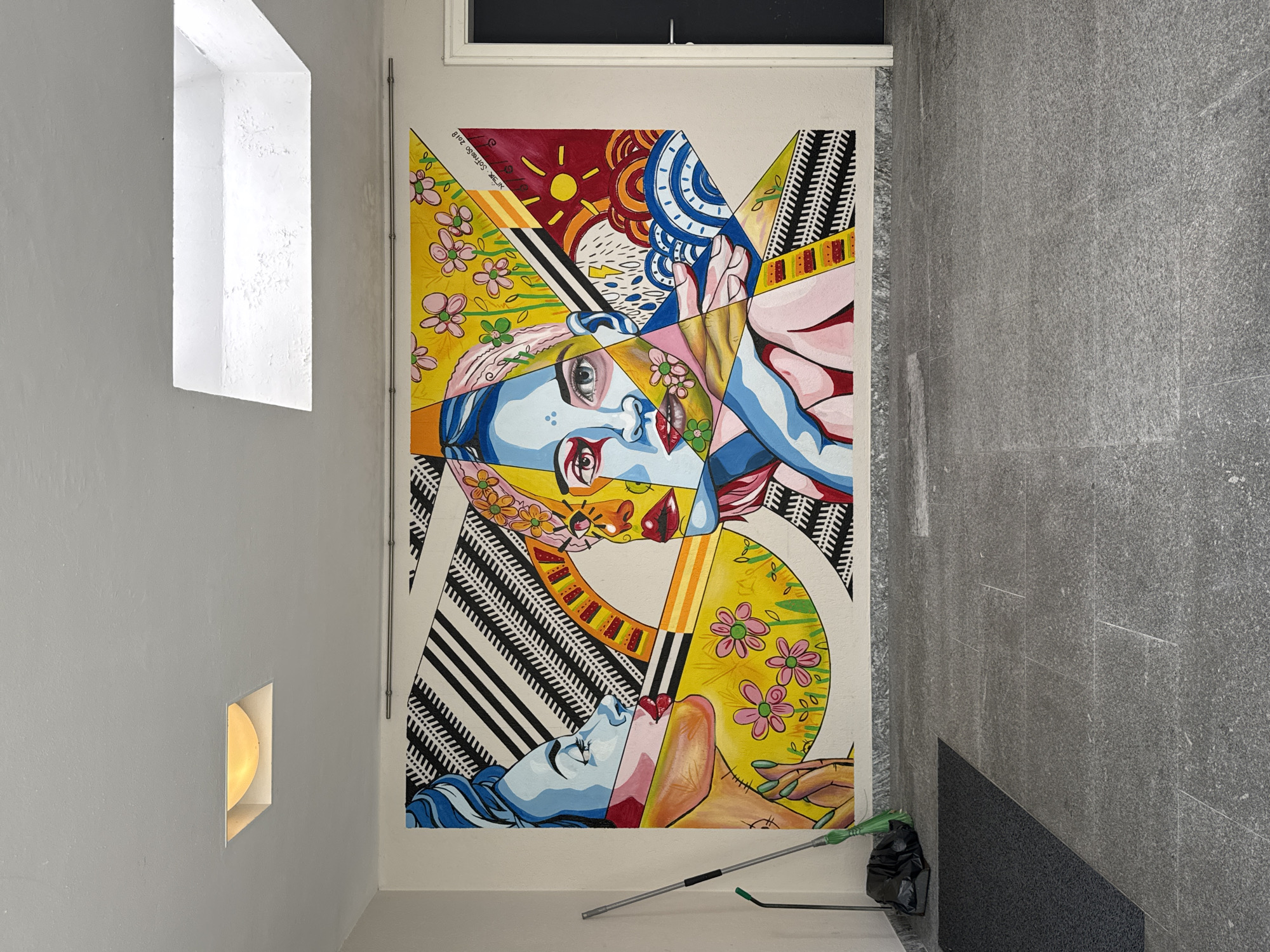 The image depicts a vibrant mural painted on a white wall, likely in an urban setting. The mural is a colorful, abstract portrayal of two faces, rendered in a bold, graphic style with sharp lines and geometric shapes. One face, on the left, is depicted in profile, with blue hair and skin, and a pink lipstick-kissed mouth. The other face, on the right, is more fragmented, with prominent blue and yellow tones, and detailed eyes. The background of the mural features abstract patterns, including black and white diagonal stripes, a semi-circular fan-like motif, and floral elements with pink and yellow flowers. The mural is signed "N'SGR Soffredo 2018".
In the foreground, the floor is covered with large, grey granite tiles. To the left of the mural, a green-handled broom with a black dustpan and a black garbage bag are placed on the floor. The wall meets the floor with a grey granite baseboard. Above the mural, the ceiling is white, with two recessed skylights, one of which is illuminated. To the right of the mural, a dark grey door with a silver handle is visible.
The location is identified as Locarno, Switzerland. This mural is located in an outdoor, covered area, possibly a public walkway or entrance, given the presence of the broom and garbage bag, and the floor tiling. The overall impression is one of a modern art installation within a functional urban space.