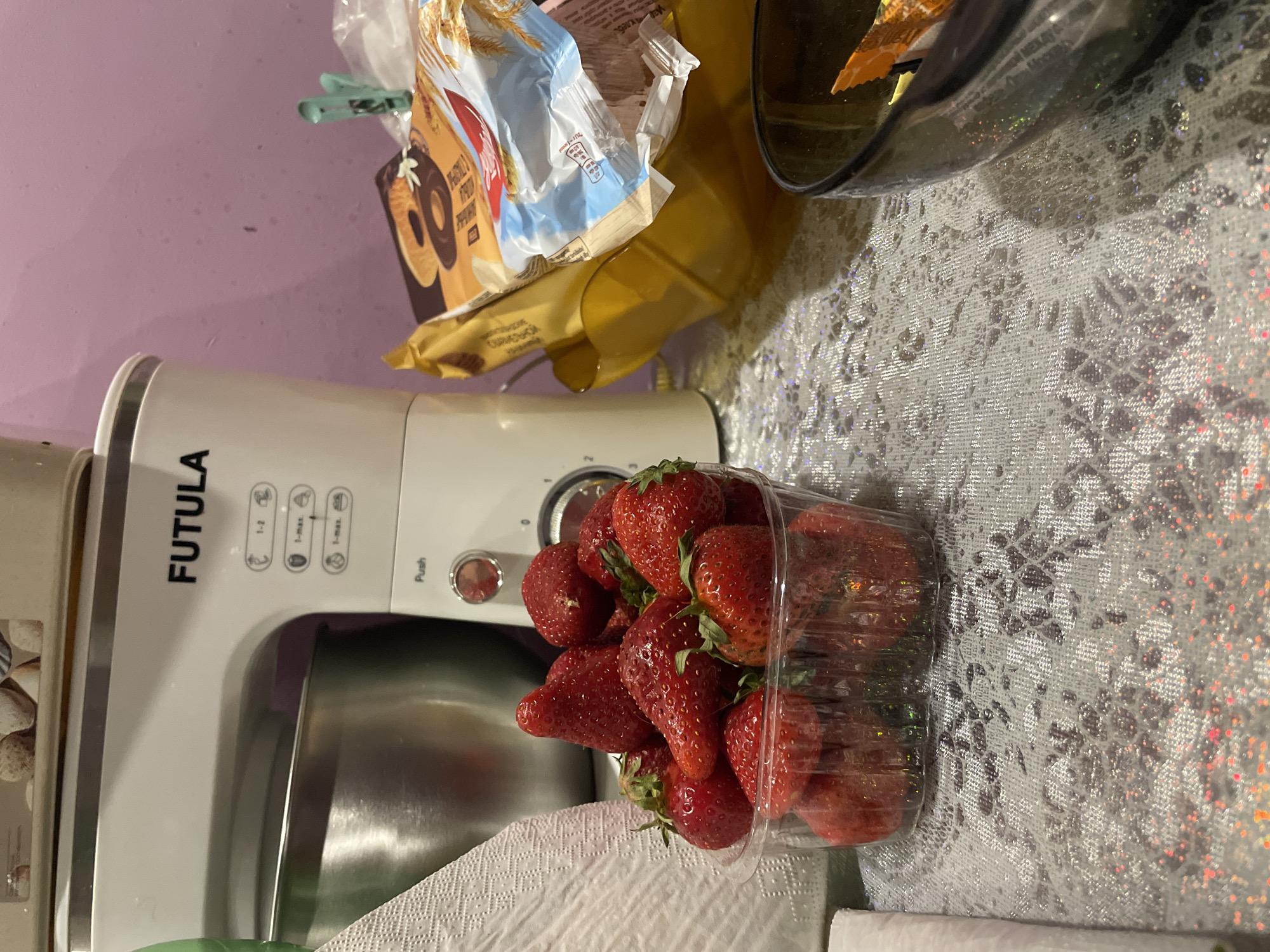 The image presents a countertop scene. A container of fresh strawberries sits prominently in the foreground, positioned on a white, textured paper towel. Behind the strawberries, a white Futula-branded stand mixer is partially visible. Further back, a package of cookies is held closed with a green clip, next to a yellow container and a dark bowl, all on a surface covered with a glittery, silver-patterned tablecloth. A portion of a lavender-colored wall is visible in the background. The scene appears to be indoors, likely in a kitchen. The location in Olginka, Russia, is inferred.
