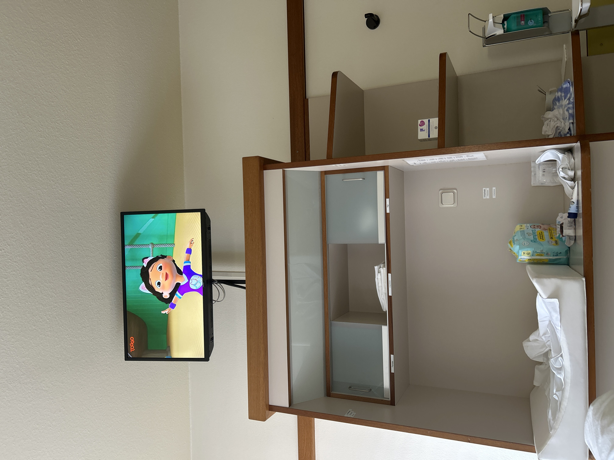 This is a full-shot, eye-level photograph of a baby changing station in a hospital room. The room appears to be in Gummersbach, Germany, based on the hospital's website. A flat-screen television is mounted on the wall above the changing station, displaying a cartoon of a young girl with cat ears. The changing station itself is a white, padded surface with white sheets. To the right of the changing station, a package of Pampers diapers is visible. Below the television, a cabinet with glass doors contains various items, including folded white cloths. The overall environment is clean and functional, indicative of a hospital setting.
