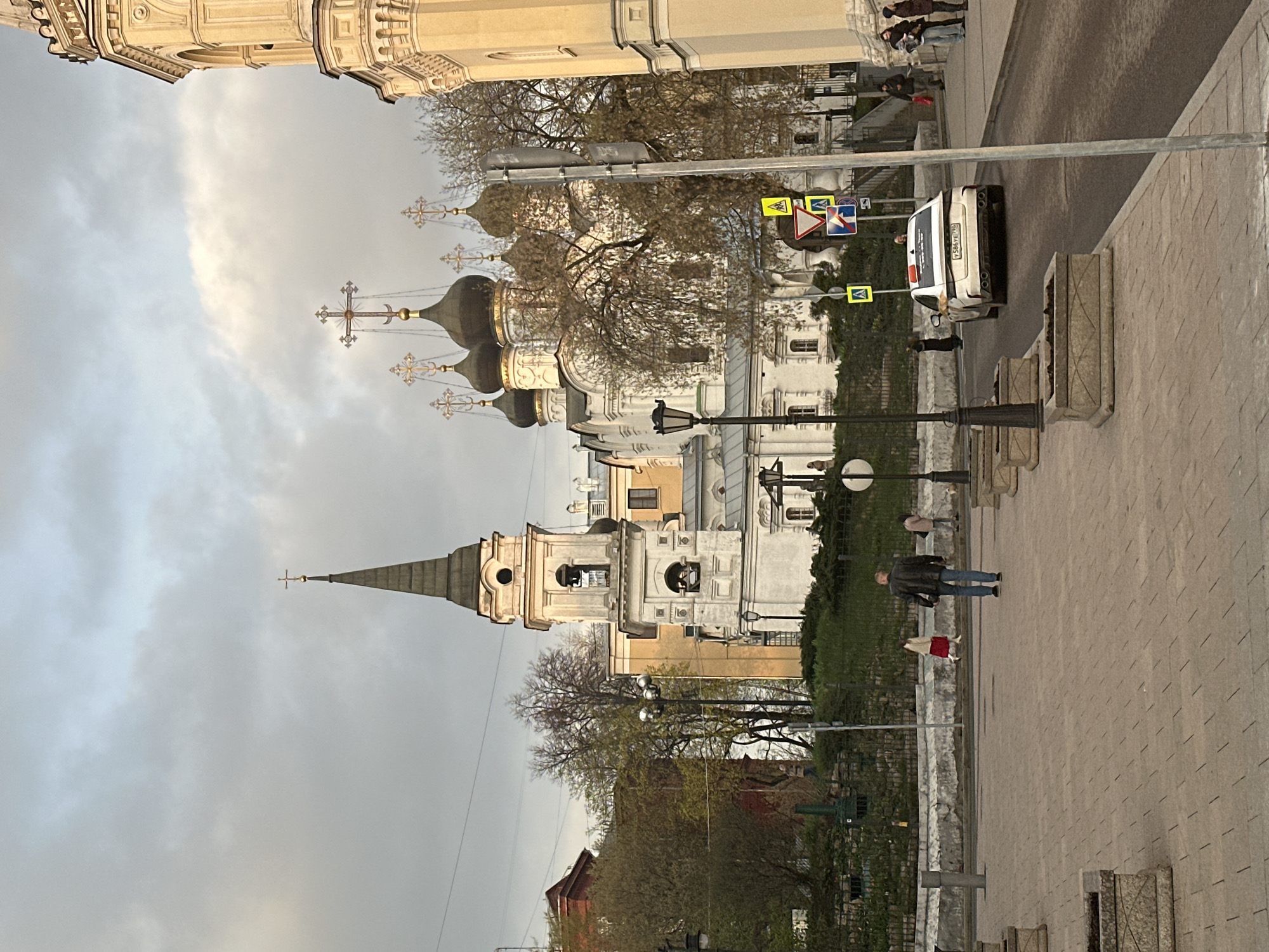 The image displays an urban street scene in Moscow, dominated by several Orthodox churches under an overcast sky, likely during late afternoon or early evening.
In the mid-ground, a prominent Orthodox church features white walls and multiple dark, onion-shaped domes, each crowned with a golden Orthodox cross. To its left, another church is visible with a tall, pointed spire and a bell tower, also featuring light-colored architecture. On the far right, a large building with classical architectural elements provides a partial backdrop.
A paved road occupies the middle-right of the frame, alongside a sidewalk. A white car is parked on the street, displaying a Russian license plate: "Y 583 YA 196 RUS." A pole nearby holds several traffic signs, including yellow diamond-shaped signs with black pedestrian symbols, a red-bordered white triangle warning for pedestrians, a blue directional sign, and a triangular yield sign.
Pedestrians are scattered throughout the scene. On the sidewalk, a person in a dark jacket and blue jeans walks away, while another in lighter attire walks towards the camera. A small group of people is also visible on the far right. Bare-branched trees are interspersed among the buildings, suggesting early spring or late autumn. Street lamps and concrete planters line the sidewalk. The diffused natural light from the cloudy sky creates soft, even illumination.