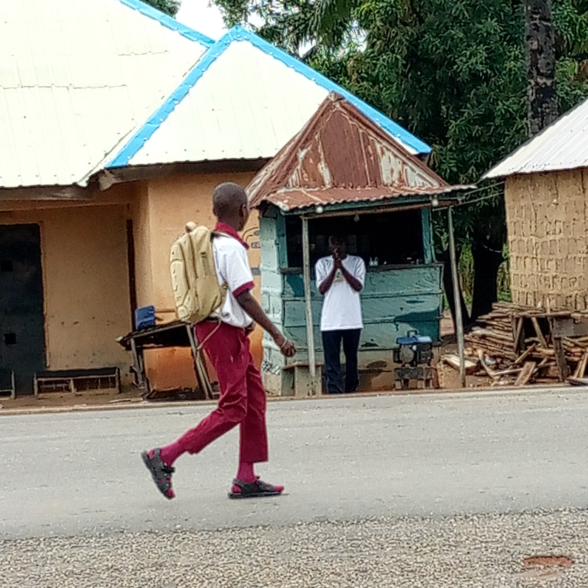 A young person wearing a white shirt with a red collar, maroon pants, maroon socks, and sandals, and carrying a beige backpack is walking on an asphalt road in Mararaba, Nigeria. Behind the person, there's a blue-green wooden kiosk with a rusted metal roof, where another person in a white t-shirt and dark pants is standing. To the left of the kiosk is a building with a white roof and blue trim, and to the right is a structure made of earth bricks and a pile of wooden planks. Green trees are visible in the background.