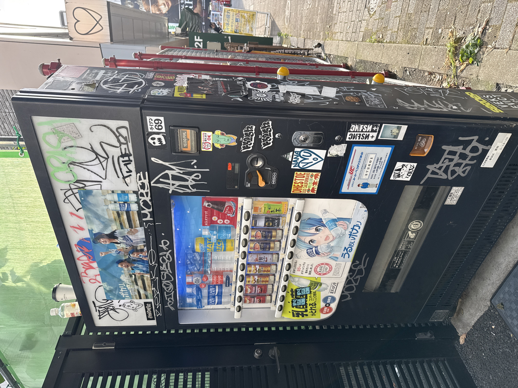 A vending machine, covered in graffiti and stickers, stands on a sidewalk in Shinjuku, Japan. The machine is black with a transparent front displaying various drinks and snacks. Several stickers with Japanese text and images are applied to the machine's exterior. The sidewalk is paved with bricks, and a metal fence with red posts runs alongside the machine. In the background, a street is visible, with buildings and a yellow sign. The overall scene suggests an urban environment.