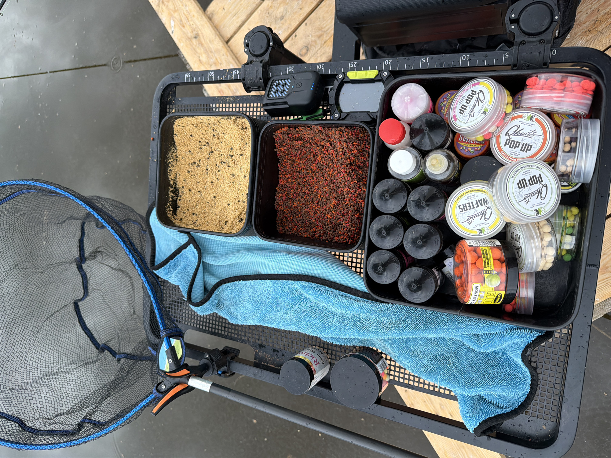 Here's a description of the image:
The image shows a fishing bait tray positioned on a damp surface, likely near water. The tray contains various types of bait, including ground bait in two square containers on the left side. One container has a light-colored bait, and the other contains a darker, red-brown bait.
To the right of the ground bait, there are numerous round containers with black lids, as well as several clear plastic containers filled with colorful boilies (fishing bait). The labels on the containers are in both English and Russian and advertise "POP UP," "WAFTERS," and other flavor varieties.
A blue towel partially covers the tray's contents, and a fishing net is visible on the left side of the tray. A rod and reel and additional bait containers are visible below the tray. The scene is outdoors, as evidenced by the moisture on the tray and the wooden planks visible in the background. There is no indication of it being in Прикамский, Russia.