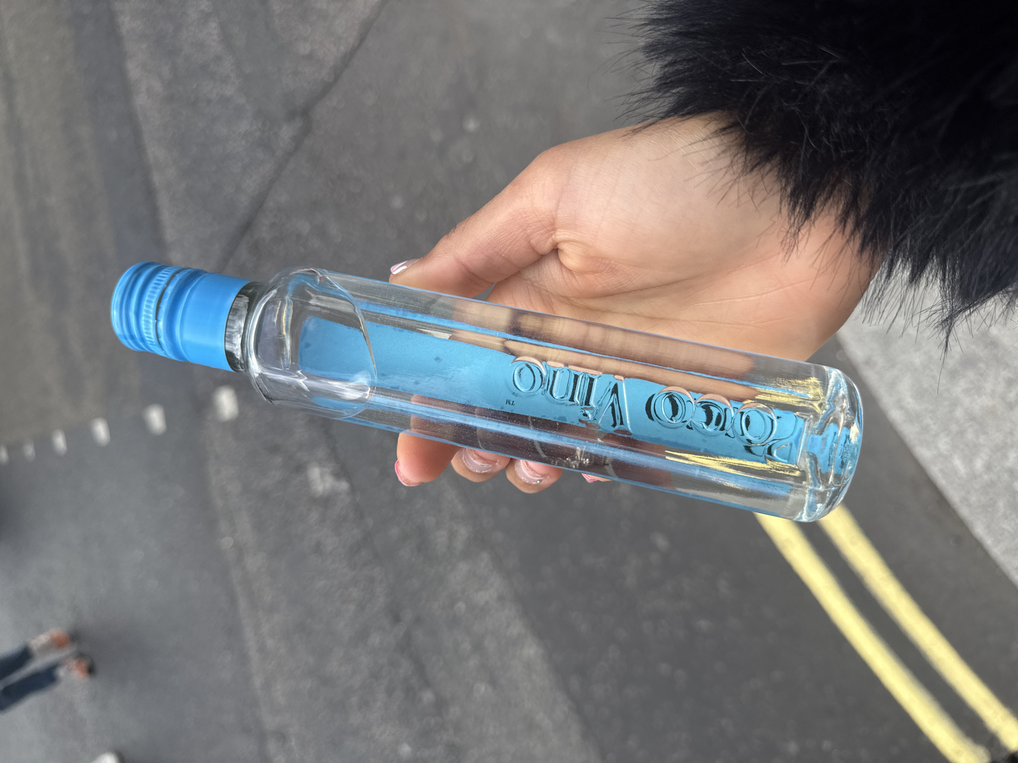 A person's hand, exhibiting a light-to-medium skin tone with light pink nail polish, holds a clear, elongated glass bottle with a blue screw cap. A fluffy black cuff or sleeve is visible around the wrist.
The bottle contains a clear liquid and features a blue label visible through the contents, with the white text "Poco Vino" and a "TM" symbol printed in reverse, indicating it's seen through the bottle. The bottle is held horizontally in the frame.
The setting is outdoors, on what appears to be an asphalt or paved street in London, United Kingdom. The ground is dark grey, showing faint white dashed lines in the upper left background and prominent double yellow lines running diagonally in the lower right background. Blurred distant elements suggest other people or objects on the ground. The lighting is bright and clear, indicative of daytime.
