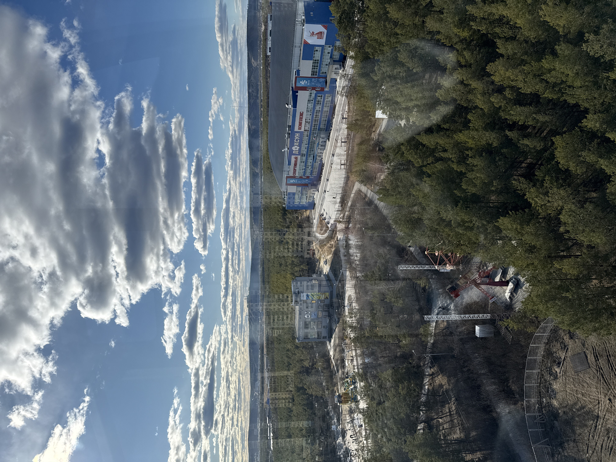 The image, likely taken from a high vantage point in Yakutsk, Russia, shows an overview of a cityscape with a reflection on a window. The sky is blue with white clouds. A building complex is visible, possibly a sports or entertainment center, with writing in Russian on its facade, including the words "Спортивный Комплекс" (Sport Complex) and "Вода" (Water). A large, curved structure is adjacent to the building complex, potentially an ice arena. A dense forest borders the building complex and the curved structure. In the foreground, a construction site is seen within the forest area. Several construction cranes are visible. The ground near the construction site appears disturbed, with traces of tire tracks and bare earth. Part of the scene seems to be overlaid with the sky reflecting in the window, making for a double exposure effect. The image shows the urban landscape interwoven with natural elements.
