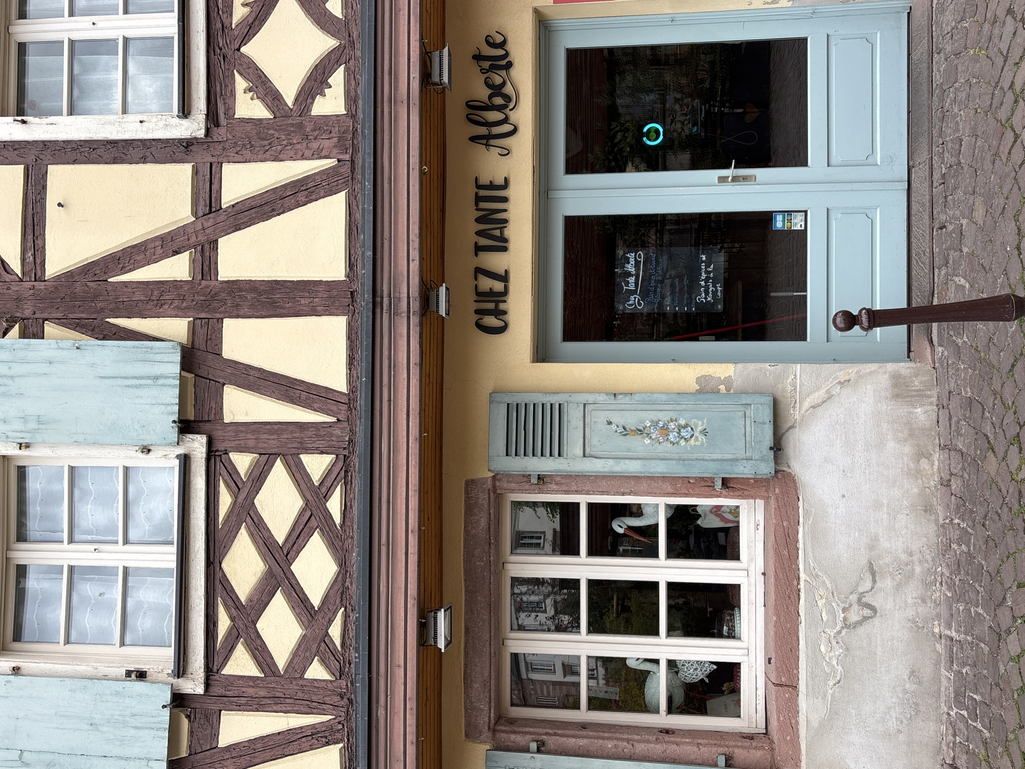 The image shows the facade of a building in Colmar, France. The building features traditional Alsatian half-timbered architecture with dark wooden beams against a light yellow stucco background. Above the storefront, a window with pale blue shutters is visible. The storefront itself has a light yellow facade with the name "CHEZ TANTE Alberte" in black lettering above a double glass door painted a light blue. To the left of the door, a window displays white stork figurines and other decorative items. The ground in front of the building is paved with cobblestones. The lighting suggests it is daytime, likely overcast.