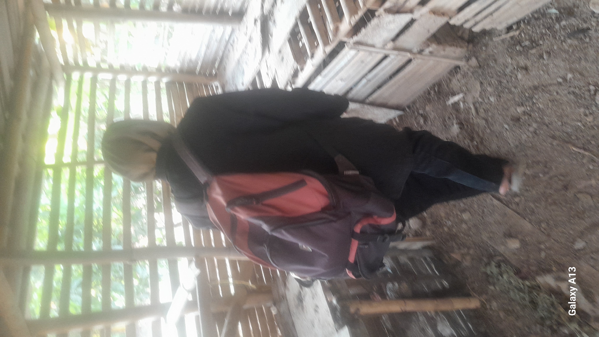 A person wearing a black jacket and dark pants, with a backpack on their back, walks away from the camera inside a wooden structure. The structure has a slatted roof and walls, and appears to be an outdoor shelter or shed. The ground is dirt and scattered debris. The person's hair is blonde and they are wearing light-colored footwear. The image also contains a watermark "Galaxy A13". The location is identified as Hadipolo, Indonesia.