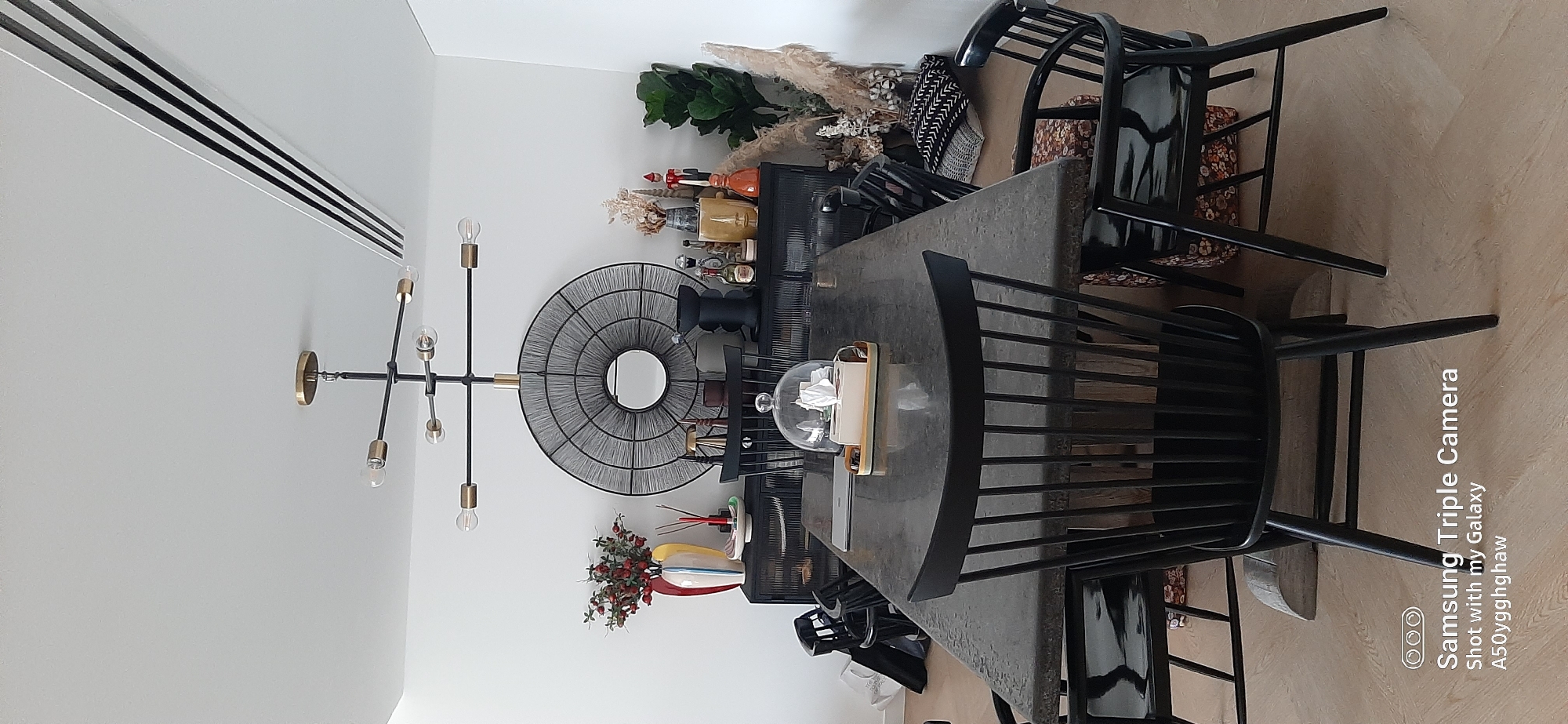 The image shows a dining room with a dark rectangular table surrounded by black chairs. A black sideboard with a textured front is against a white wall, adorned with decorative items including a round, textured mirror, a vase with flowers, and various bottles and figurines. A modern chandelier hangs from the ceiling. The flooring is light-colored wood. The scene is clean and organized, suggesting a well-maintained residential interior. The "Samsung Triple Camera Shot with my Galaxy A50yggghaw" watermark in the lower left corner indicates the image was captured by a Samsung smartphone. There is no indication of the location being Dubai, United Arab Emirates.