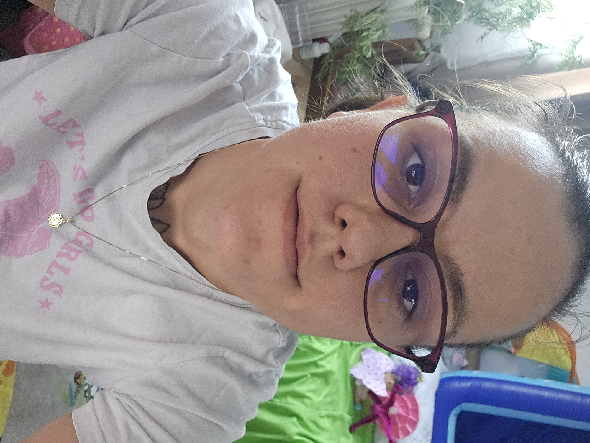 The image is a close-up selfie of a person wearing glasses and a light-colored t-shirt with pink lettering and graphics. The person has dark hair pulled back. In the background, there are some plants to the left, and to the right, there is a green draped surface, a blue inflatable structure, and other colorful objects, possibly toys. The scene appears to be indoors, likely a domestic setting, but the specific location in Sofia, Bulgaria, is not determinable from the visual information provided.