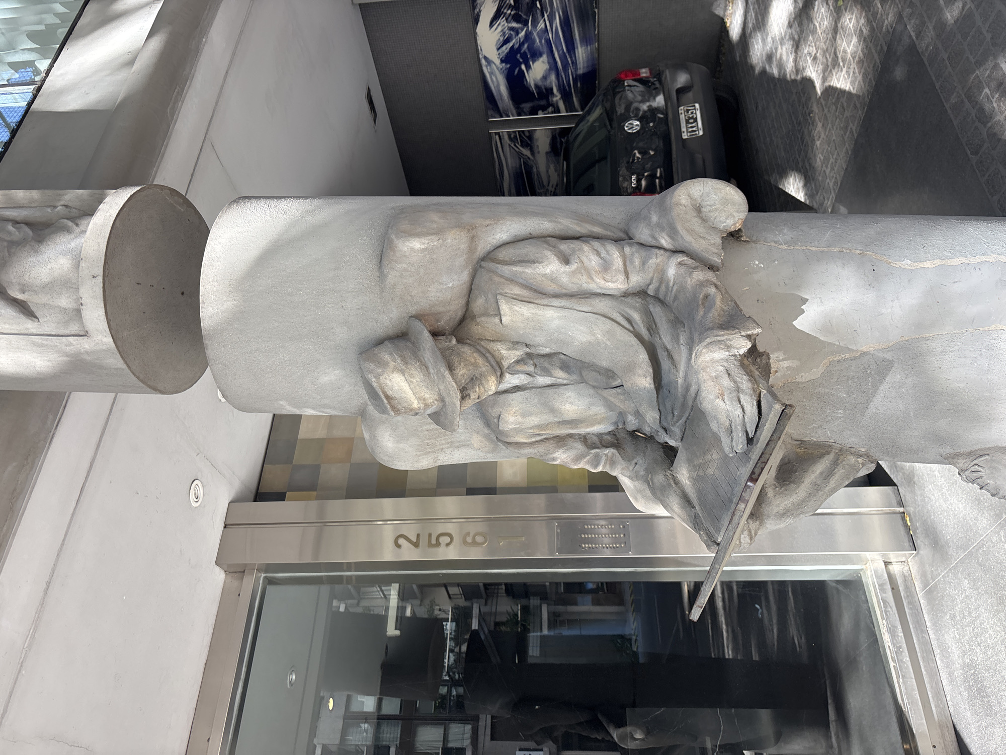The image captures a vertical outdoor scene, prominently featuring a large, grey concrete sculpture of a seated man. The sculpture is integrated into a building's architectural column or facade. The man is depicted wearing a hat and what appears to be a long coat or robes, with his head tilted downwards, concentrating on a flat, board-like object held in his hands, suggesting he is writing, drawing, or engaged in a similar task. The sculpture has a rough, textured finish, giving it a weathered appearance.
Below the sculpture, a modern building entrance is visible, characterized by large glass doors and sleek metal framing. The building number "259" is embossed on a metallic panel directly above the entrance. Reflections in the glass hint at interior elements or the opposite side of the street.
In the background, at street level, a black Volkswagen Gol car is parked. Its rear and license plate, "IXX 357," are clearly visible. The setting is an urban environment, specifically a street in Buenos Aires, Argentina. The scene is brightly lit by natural daylight, indicated by sharp shadows. The overall composition juxtaposes traditional sculptural art with modern architecture and an active streetscape.