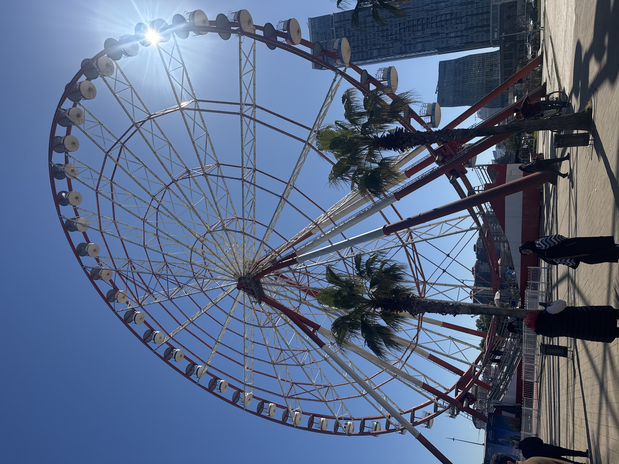 The image depicts a Ferris wheel in Batumi, Georgia. The wheel is red and white, with enclosed passenger cars. Two palm trees are positioned between the wheel's base and the passenger cars, partially obscuring the structure. Several people are visible on the ground near the wheel's base, dressed in casual clothing and some are wearing head coverings. Behind the Ferris wheel, modern high-rise buildings are seen in the background, set against a clear blue sky with a bright sun glare visible at the top of the wheel. The scene appears to be captured during daylight hours. The ground surrounding the wheel is paved and has some shadow cast across the surface.