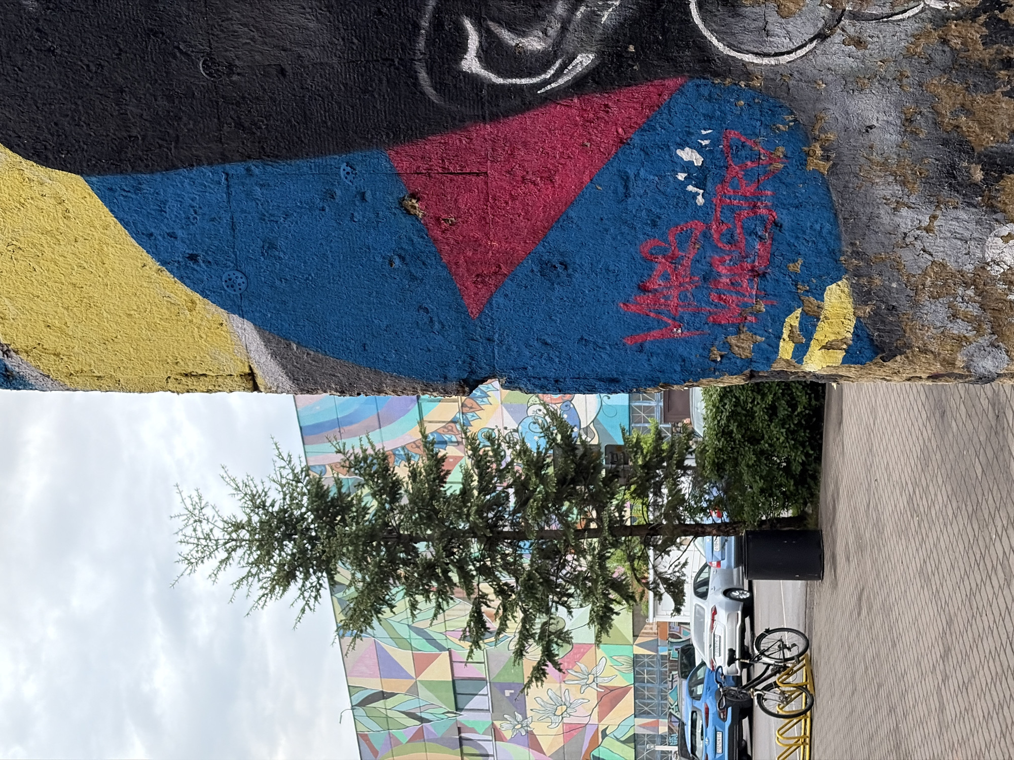 Here's a detailed description of the image:
**Overall Scene:** The image shows a split view, with the upper half depicting a section of graffiti-covered wall and the lower half revealing a cityscape featuring a painted building, a tree, parked vehicles, and a sidewalk.
**Upper Graffiti Wall:** A section of the wall displays a variety of graffiti in colors like yellow, black, blue, and red. The colors and textures appear rough and layered.
**Lower Cityscape:**
* **Building:** A colorful building with a large-scale geometric mural is visible.
* **Tree:** A thin tree stands in front of the mural.
* **Vehicles:** Parked cars are lined up along the curb, showing a mix of blue and white vehicles.
* **Sidewalk:** A brick sidewalk occupies the right side of the lower half, with a bike rack holding several bikes.
**Location:** Unknown, Unknown.