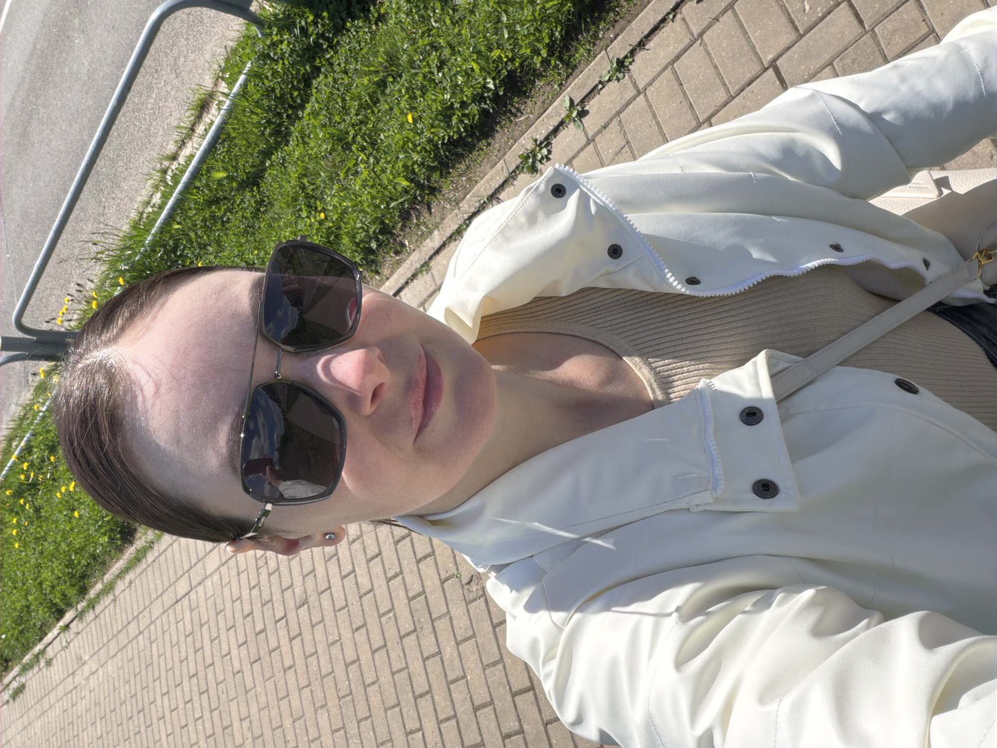 A young woman wearing sunglasses and a white jacket is standing on a brick sidewalk next to a patch of grass and a metal railing. The city is unknown. The woman is looking at the camera. The surroundings are sunny.