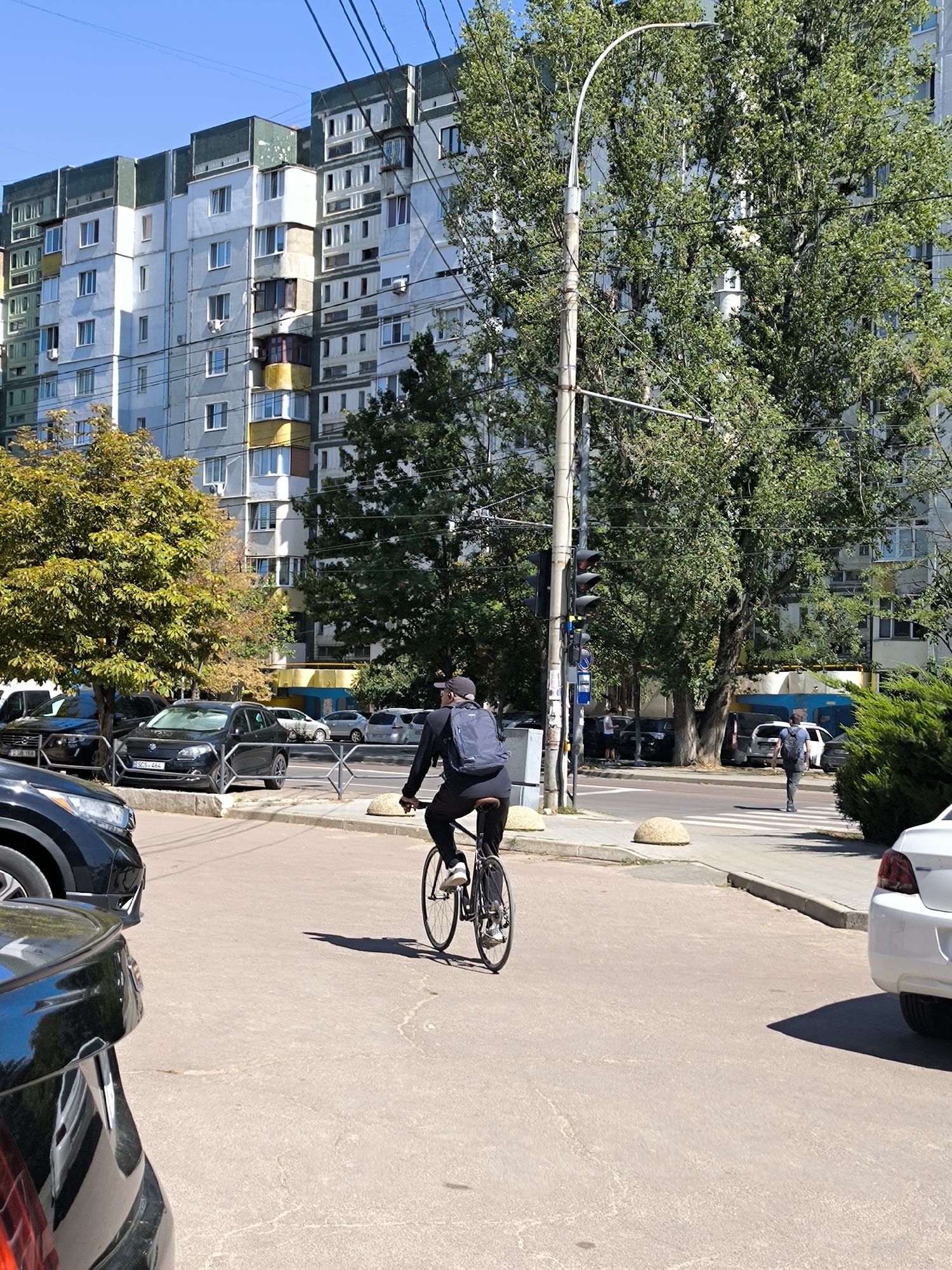 In Chișinău, Moldova, on a sunny day, a person wearing a black baseball cap and a black backpack rides a bicycle in the foreground, facing away from the camera. The cyclist is on a paved road with a white pedestrian crossing visible in the middle distance. Behind the cyclist, a tall, multi-story apartment building with a mix of white, gray, and green facades stands under a clear blue sky. Several cars are parked or driving on the road. Trees with green and yellow foliage are scattered throughout the scene, with a prominent streetlight on the right. In the background, another person walks across the pedestrian crossing, also with a backpack. The environment appears to be an urban residential area with dense housing and traffic.