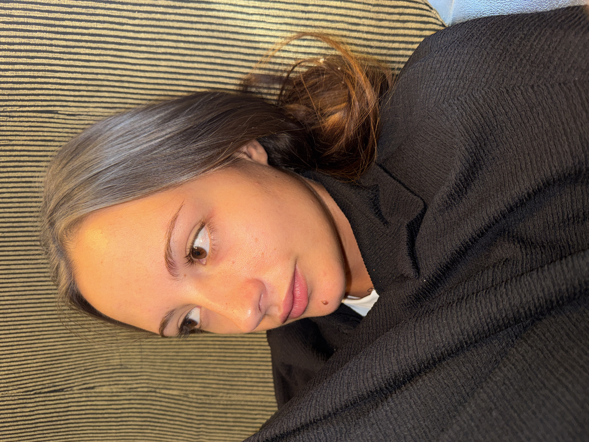 A young woman with fair skin, brown eyes, and brown hair with greyish undertones is shown in a close-up, side-profile shot. She is lying down, her head resting on a surface with a textured background of alternating dark and light vertical stripes. She is wearing a black textured garment that covers her chest and shoulder. A light-colored garment is visible underneath the black one. Her gaze is directed to her left, away from the camera. She has a mole on her chin. The scene appears to be indoors, possibly in Skala Marion, Greece, given the reference.