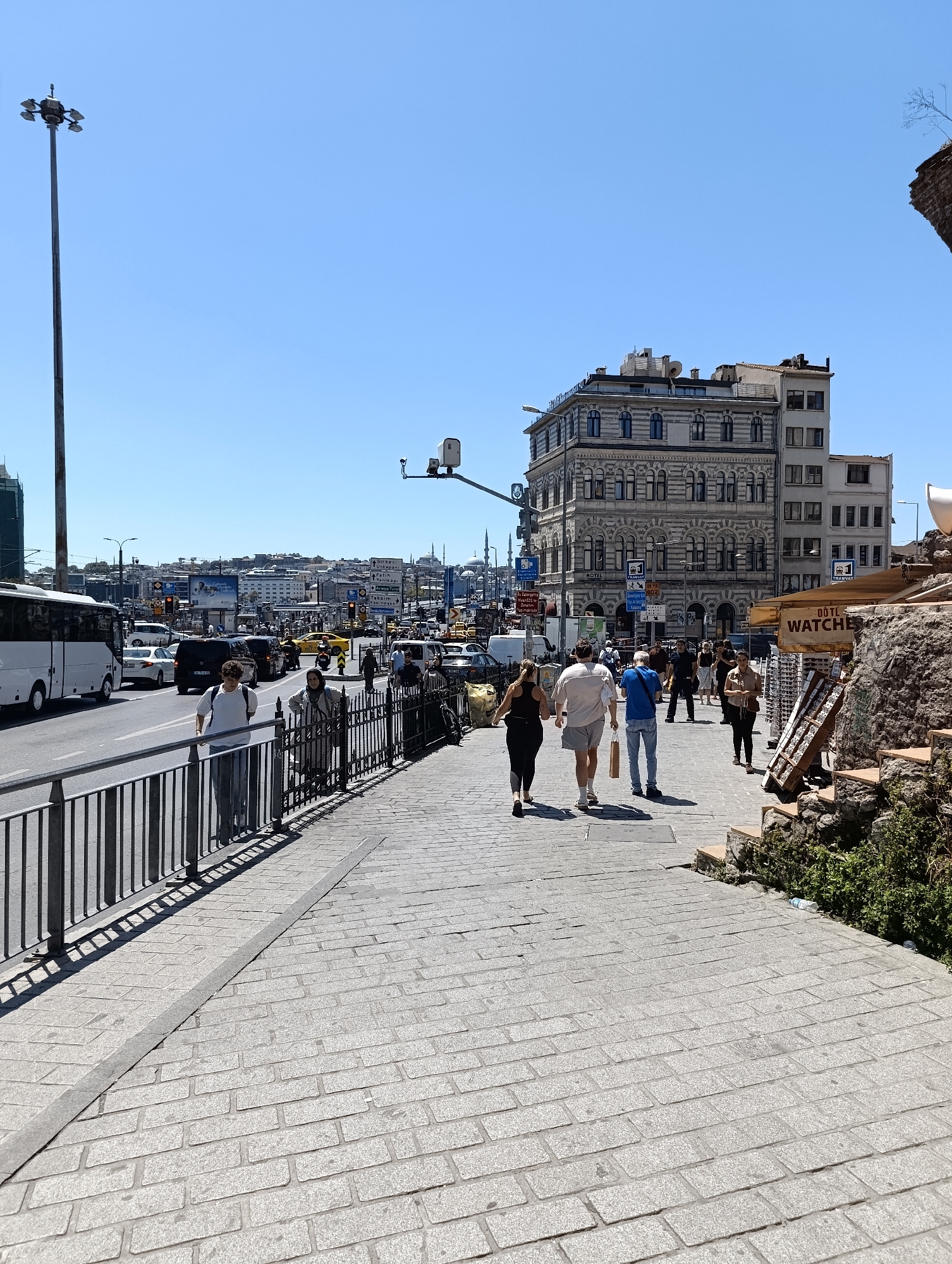 This media file depicts a sunny day scene on a pedestrian walkway in Beyoğlu, Turkey. The walkway is paved with stone pavers and bordered by a metal railing on one side, separating it from a busy street with vehicular traffic. People are walking along the sidewalk, some singly and some in small groups.
On the street, various vehicles are visible, including a white bus, cars, and yellow taxis, suggesting moderate traffic. In the background, several buildings of varying architectural styles are visible, including a prominent, ornate, multi-story building with arched windows and decorative facade. Beyond the immediate buildings, a cityscape stretches into the distance under a clear blue sky.
To the right of the walkway, there is a display of watches under a shaded awning, with some stacked on wooden racks. Adjacent to the display, a rocky, somewhat overgrown area with some stone steps suggests a more rustic or historical element.
Notable details include the bright sunlight casting shadows, indicating it is likely daytime, perhaps late morning or early afternoon. The clear sky points to fair weather. The presence of Turkish signage, such as on the traffic direction signs and the "ÖtüL WATCHES" sign, confirms the location in Turkey. The general atmosphere appears lively and active due to the presence of pedestrians and traffic.