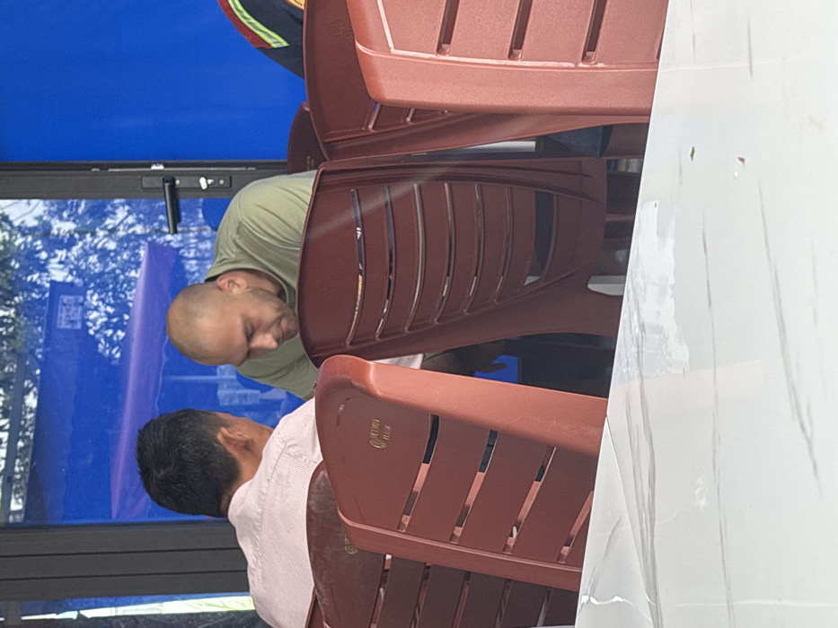 The scene depicts two individuals seated at a table within what appears to be a casual dining establishment in Ngũ Hành Sơn Ward, Vietnam. In the foreground, a white table with subtle grey veining is visible. Several maroon-red plastic chairs with horizontal slat backs are arranged around the table.
Two men are the primary subjects. The individual on the left is seen from the back, possessing short, dark hair and wearing a light pink or off-white collared shirt. He is oriented towards the right side of the frame. The individual on the right is a Caucasian male with a bald head and a short beard, wearing an olive green crew-neck t-shirt. He is seen in profile, looking downwards, with his body slightly angled towards the first man, suggesting an interaction or shared focus.
The background features a large window or glass panel, reflecting a vibrant blue element, possibly an awning or wall, along with blurred green foliage. A black door frame with a black handle is visible to the right of the bald man. The lighting suggests daytime. No other specific objects or actions are clearly identifiable.