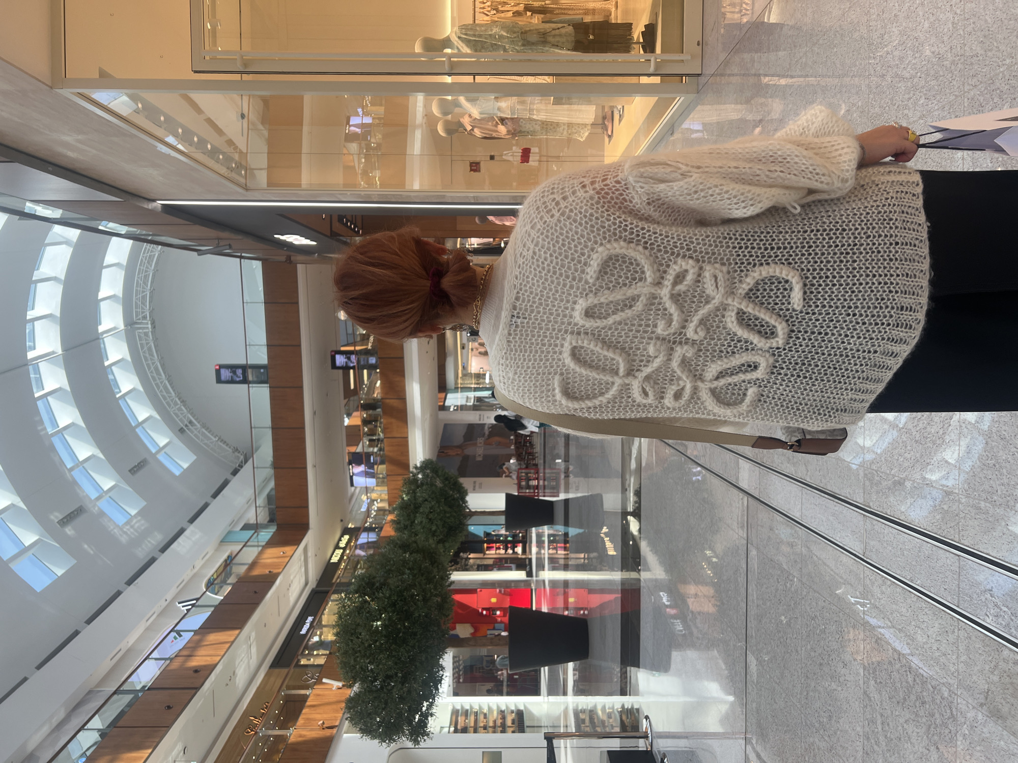 The photograph depicts a woman standing in a shopping mall in Dubai, United Arab Emirates. The woman has reddish-brown hair pulled back in a bun. She wears a cream-colored knitted cardigan with a prominent logo pattern on the back. She is holding shopping bags. The mall is modern, featuring multiple levels with storefronts and escalators. There are trees in planters along the mall's walkway. A clear domed ceiling with numerous rectangular windows allows natural light to illuminate the interior of the mall. The storefront windows showcase fashion displays with mannequins. The floor is light gray and polished.