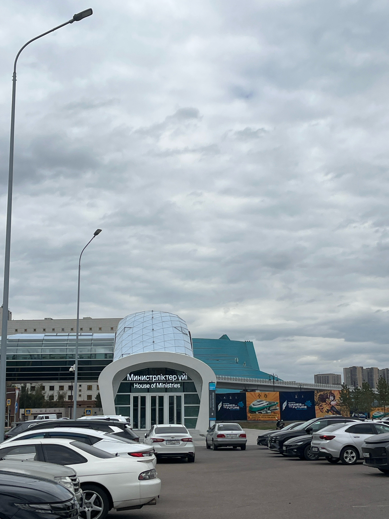 The image shows a cloudy day in Astana, Kazakhstan, with the "House of Ministries" building in the background. The building features modern architecture with a glass dome and teal-colored roof. In front of the building is a parking lot with several cars, including white sedans and darker-colored SUVs. A tall street lamp is visible on the left side of the frame. There are banners in the background advertising "Games of the Future". No people are clearly visible.