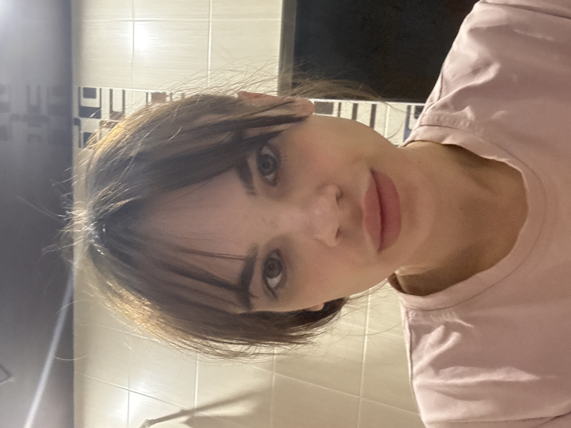 The image is a close-up, rotated portrait of a young woman looking directly at the camera. Her head is tilted slightly to her right, with her dark hair, featuring subtle lighter brown or auburn highlights, framing her face. She has bangs across her forehead, and some individual hair strands are visible, giving her a casual appearance. Her eyes are dark, likely brown or hazel, and she maintains a neutral, calm expression with her lips closed. She is wearing a light pink, possibly cotton, t-shirt.
The background consists of light-colored, likely cream or white, rectangular wall tiles. Some sections of the tiled wall feature a repeating geometric pattern in dark gray or black, creating an abstract design. To the left and upper left of the frame, there is a darker, shadowed area, possibly another wall or a piece of furniture. The lighting in the scene appears to be artificial, casting soft reflections on the tiled surfaces.
The woman is posing for a photograph, likely a selfie, given the direct gaze and close-up composition. Her posture suggests a relaxed state. No text is visible in the image. The provided context indicates the location is Rostov-on-Don, Russia, though no specific visual cues to this location are present in the image itself. The original image is oriented 90 degrees clockwise, with the woman's head on the right side of the frame and her body extending towards the bottom.