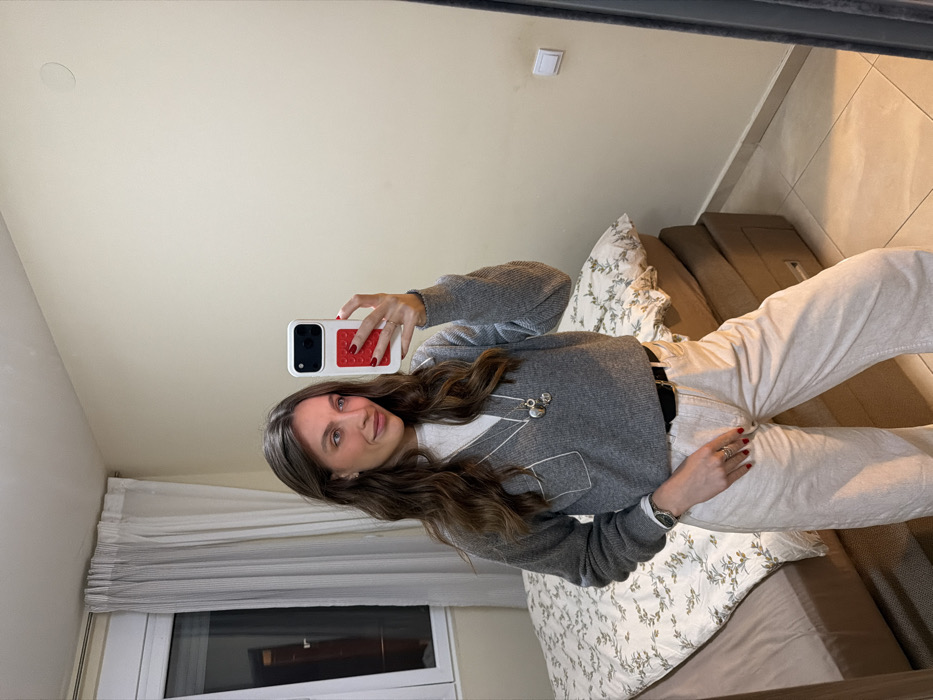 A woman stands in front of a mirror taking a selfie with her phone, which has a red textured case. She wears a grey sweater with white detailing, light-colored jeans with a black belt, and a watch. Her hair is long and wavy. The background shows a bed with patterned pillows, part of a window with white curtains, and a beige wall with a light switch. The floor appears to be tiled.