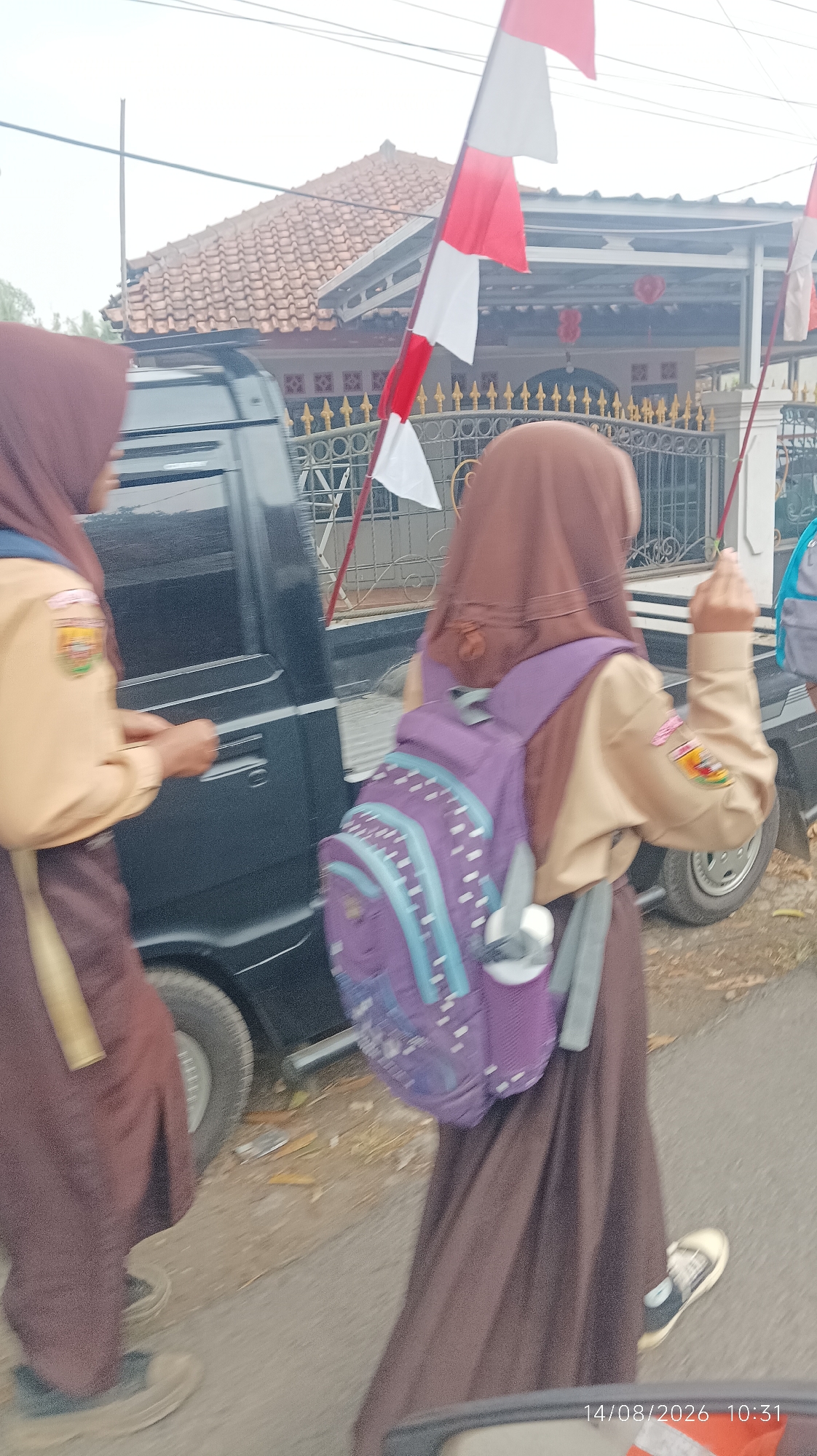 Two young girls wearing tan school uniforms with brown headscarves and brown skirts walk on a paved road next to a dark-colored pickup truck. The girl on the left is partially visible, facing the side, and holding a door handle of the truck. She has a badge on her left sleeve. The girl on the right is walking away from the camera, carrying a purple backpack with a white and light blue polka dot pattern. She also has a badge on her left sleeve. Red and white Indonesian flags are visible in the background, attached to poles. A building with a red-tiled roof and ornate fencing is situated behind the flags. The scene appears to be a street in a residential area in Tasikmalaya, Indonesia. The timestamp "14/08/2026 10:31" is visible in the lower right corner.
