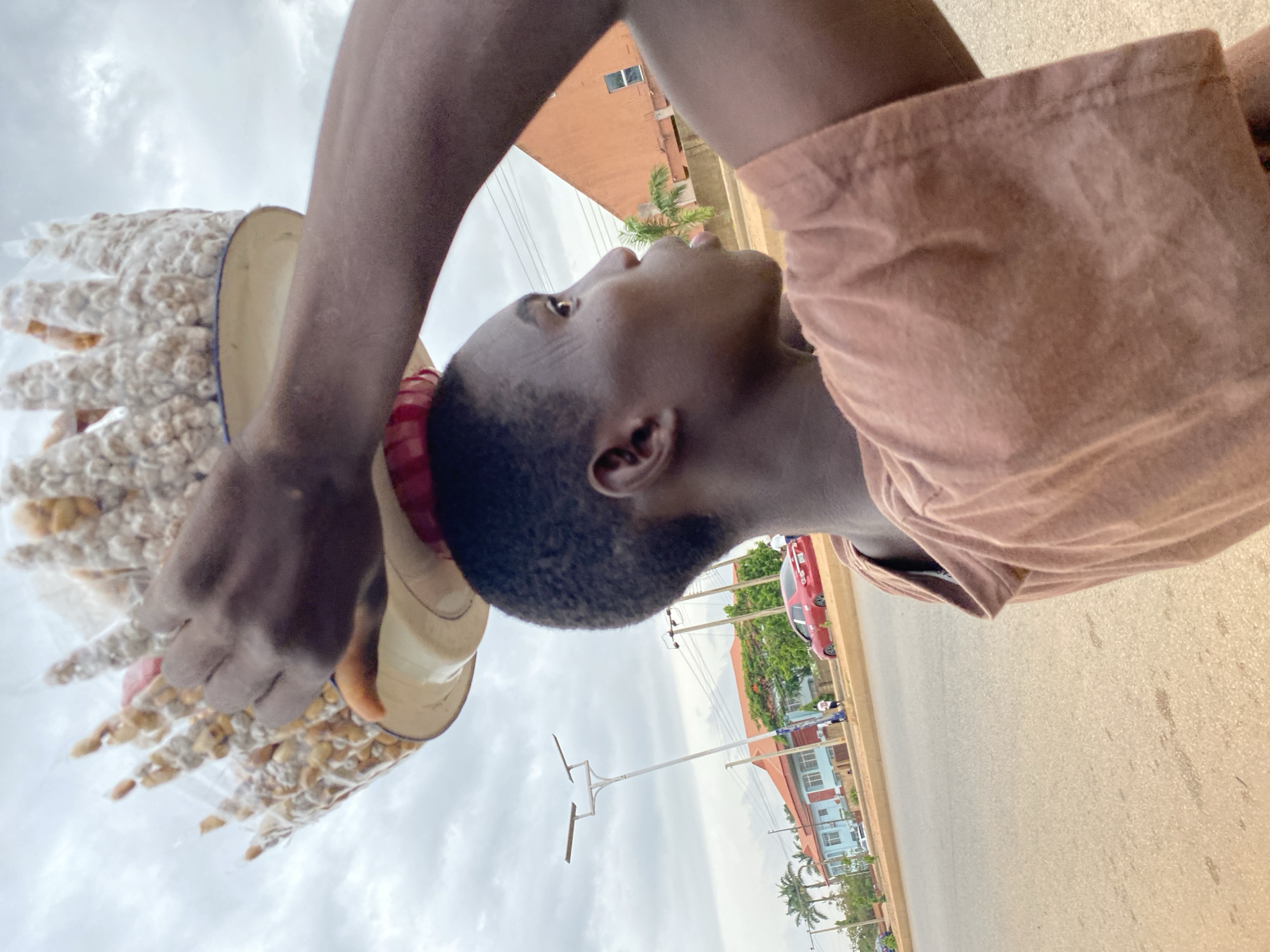 A street scene captured from a low angle features a dark-skinned male vendor in Bukuru, Nigeria. He is carrying a yellow tray filled with small, pre-packaged goods, possibly snacks or nuts, sealed in clear plastic bags. He wears a salmon-colored short-sleeved shirt and has a red wristband. In the background, the asphalt road stretches out, flanked by buildings. There's a red car parked by the roadside. Palm trees and other greenery suggest a tropical environment, and utility poles are visible against a cloudy sky.