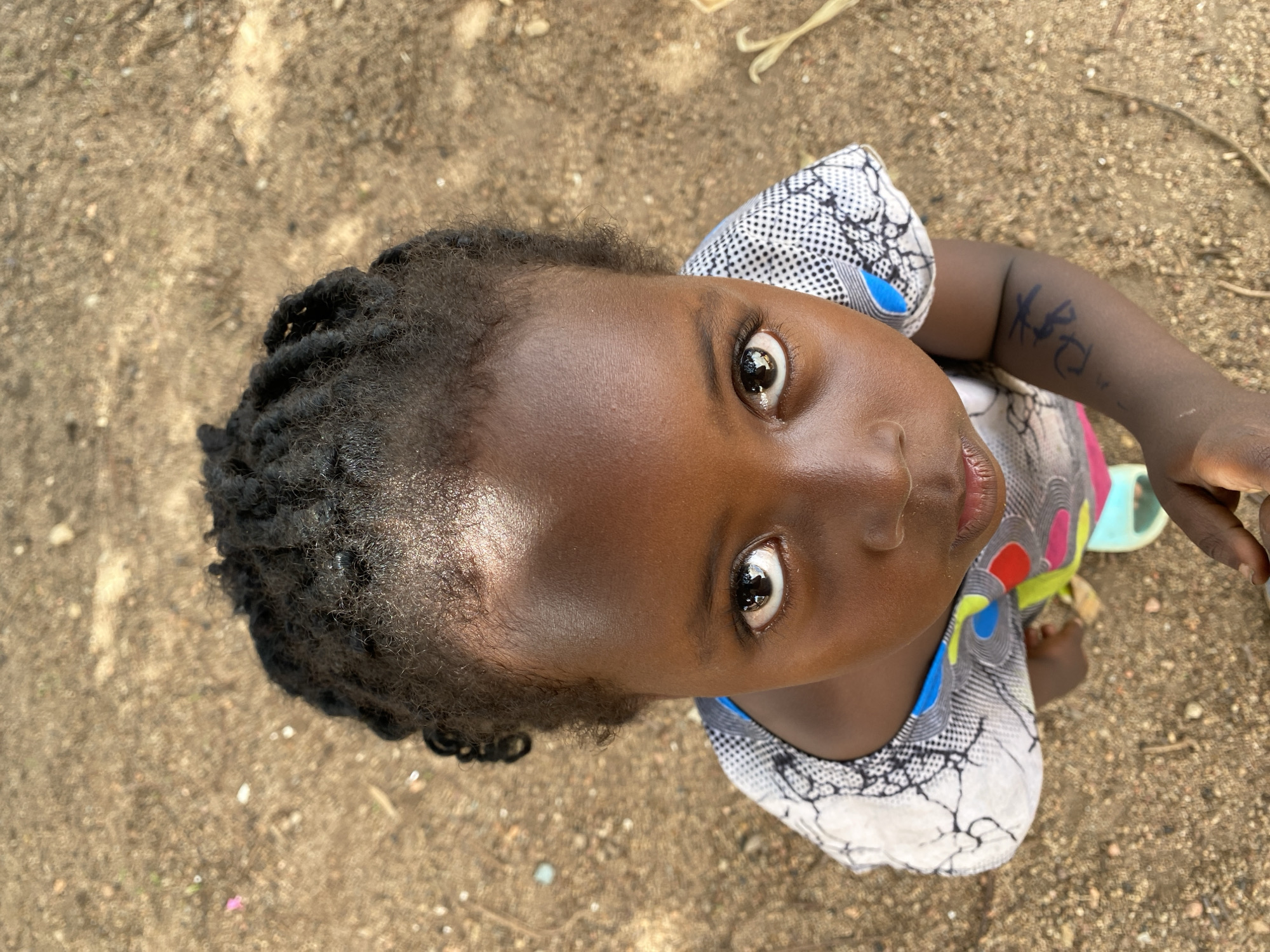 A high-angle, close-up shot features a young Black child looking directly upwards at the camera. The child has dark skin, wide, expressive brown eyes, and black hair styled in short dreadlocks or braids. Her gaze is intense and direct, suggesting curiosity or engagement with the photographer.
She is wearing a patterned garment, possibly a top or a dress, which features a mix of white, black, blue, red, and yellow geometric and abstract designs. The background is a sandy or dirt ground, hinting at an outdoor setting. The bright lighting indicates it is daytime, possibly sunny, with reflections visible in the child's eyes.
Notable details include slightly parted lips and a handwritten marking on her right arm (viewer's left), which appears to be black lettering resembling "KBC" or similar simple shapes. A light blue or green sandal is partially visible on the ground near her right side. The scene is set in Kuru Dungus, Nigeria.