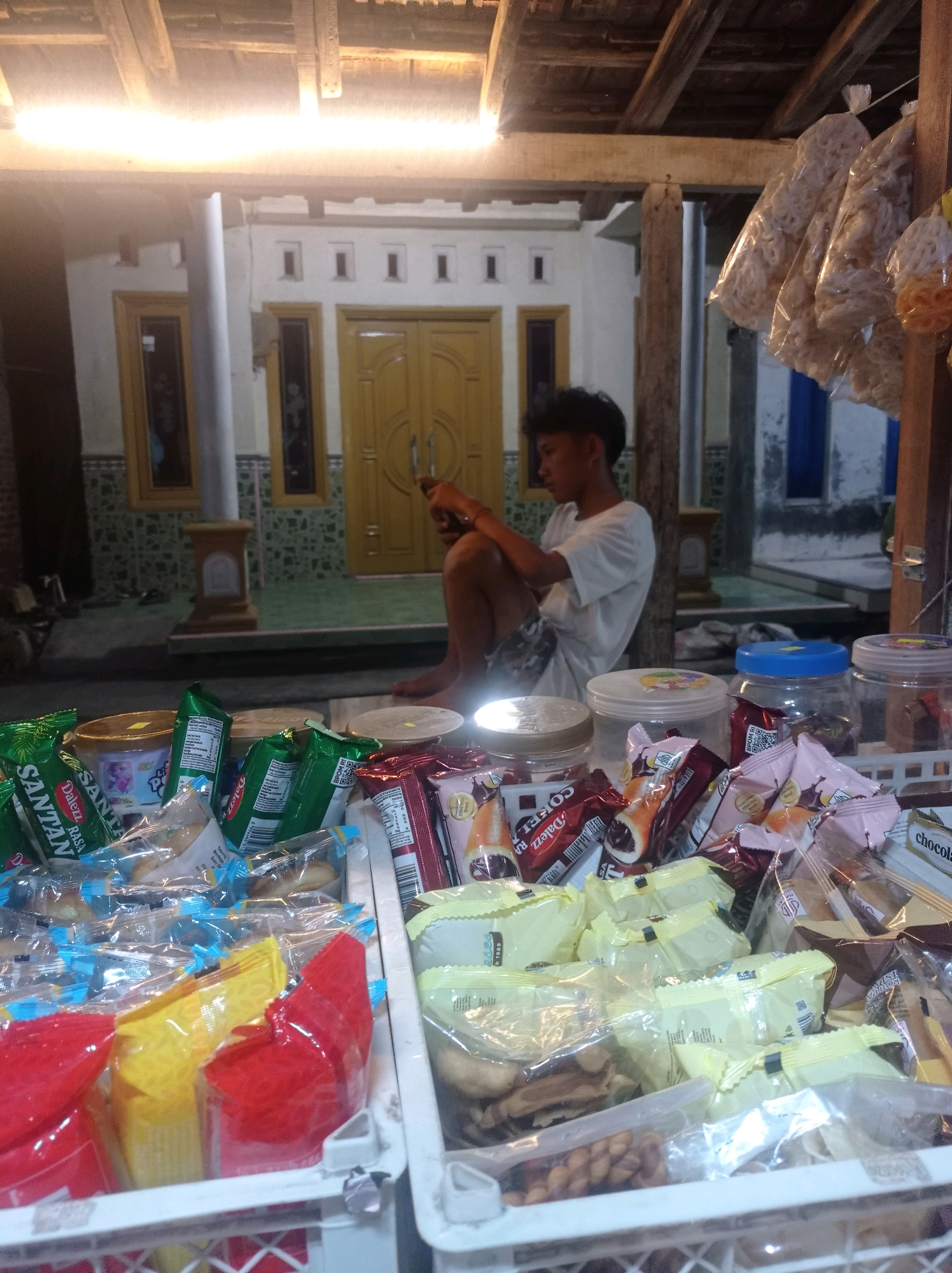 In Babalan, Indonesia, a young man sits on a raised platform in front of a building, engrossed in his phone. The building features a yellow double door and windows, suggesting a residential or commercial establishment. A covered wooden structure overhead, possibly a porch or awning, provides shade. To the right, a display of snacks hangs in transparent plastic bags, suggesting a small roadside stall or shop. In the foreground, various packaged snacks, including brightly colored wrappers, are arranged in white plastic crates, implying a retail setting where these items are sold. The scene is illuminated by artificial light, indicating it is likely evening or night.