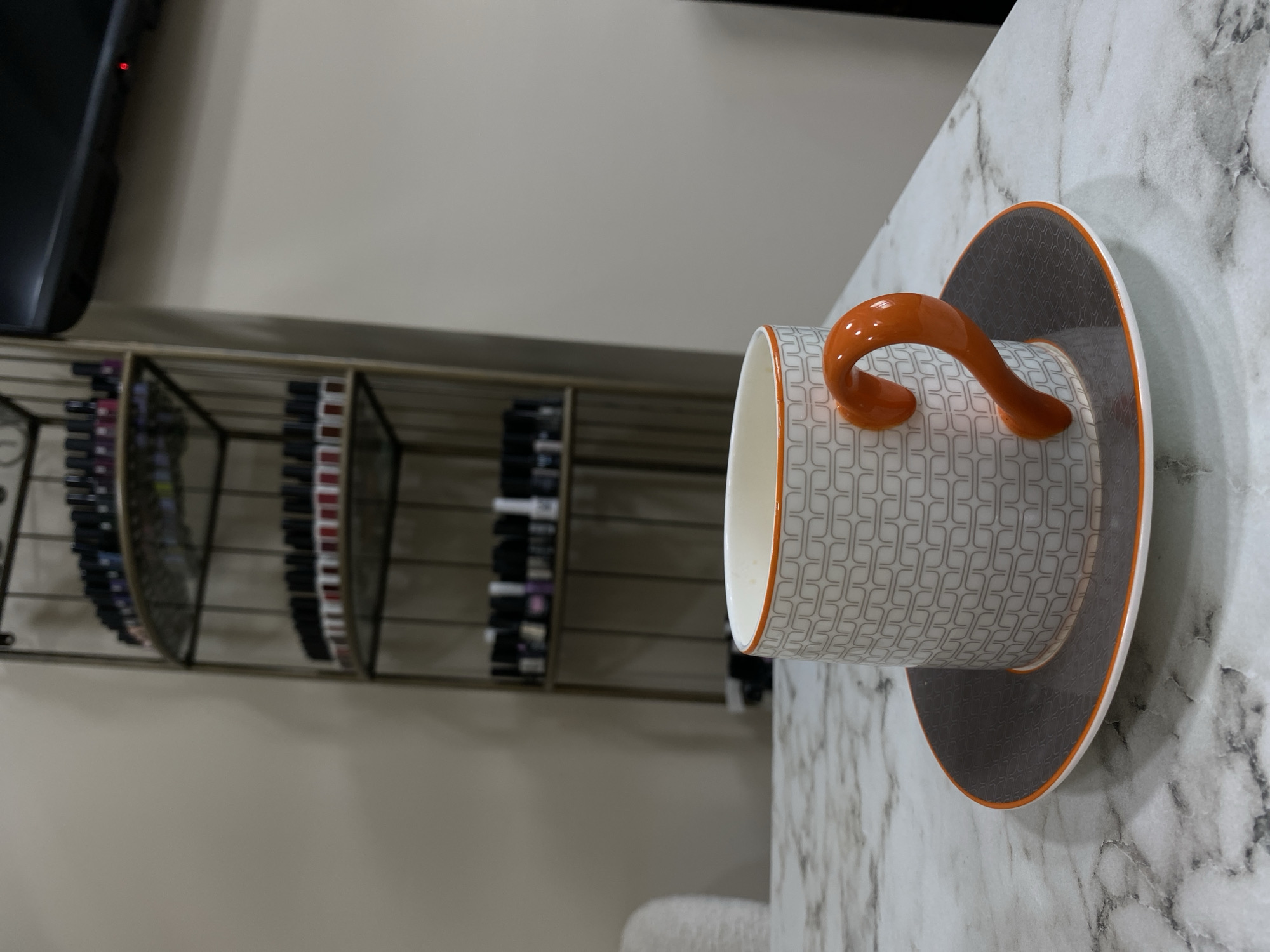 A teacup and saucer are prominently positioned on a marble-patterned surface, with the teacup featuring a white background and gray geometric pattern, accented by an orange handle and trim. In the background, there's a metal shelving unit holding numerous bottles, likely nail polish, which is typical for a salon environment. To the side of the shelving unit, a television is mounted on the wall. The setting appears to be indoors, potentially in a salon or a private residence that has salon-like setup. There are no visible indications to pinpoint the location to Sochi, Russia, beyond the general aesthetics of modern interiors.