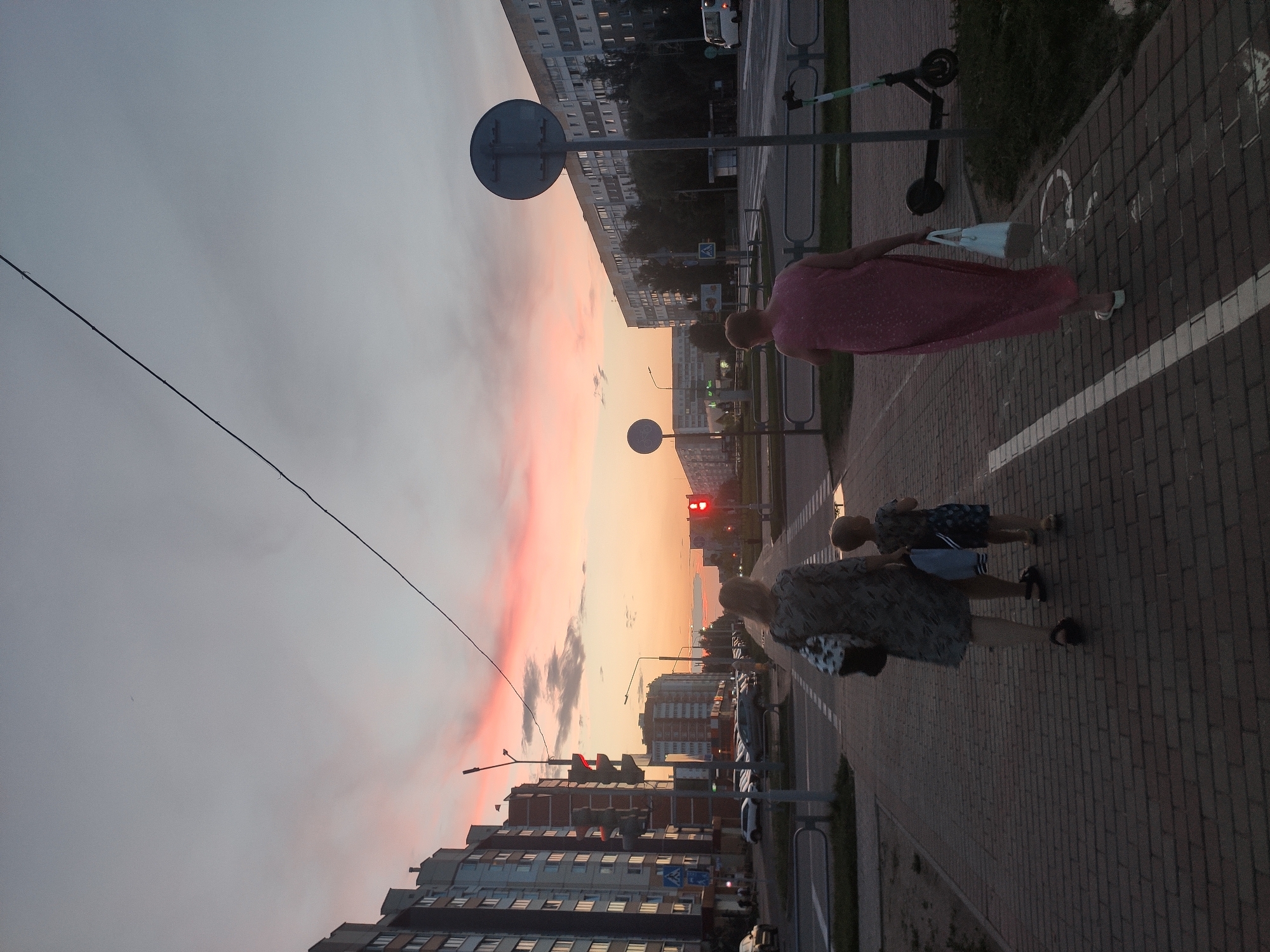 This is a full-frame, eye-level, vertical shot of a city street in Navapolatsk, Belarus during twilight. The sky exhibits a vibrant sunset with streaks of pink and orange clouds. In the foreground, a brick-paved sidewalk runs parallel to a street lined with apartment buildings.
Two women and a child are walking away from the camera on the sidewalk. The woman on the left is wearing a patterned dress and carrying a bag. The child, to her right, is wearing a dark blue patterned t-shirt and shorts. The woman on the right is wearing a long, light pink dress and carrying a white tote bag.
Across the street, traffic lights are visible, with one showing a red signal. Various vehicles, including cars and a white van, are parked or driving on the street. A scooter is parked on the grassy area next to the sidewalk. Streetlights and traffic signs are also present. The overall scene captures a common urban evening atmosphere.