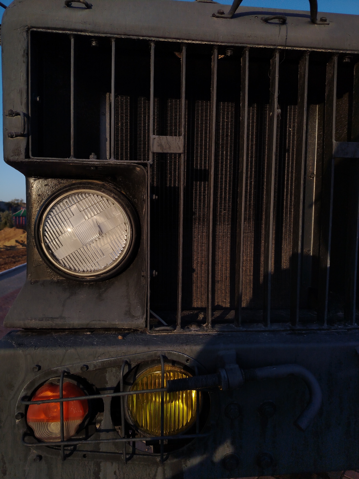 This close-up, vertical, eye-level shot features the front of a heavy-duty, dark gray truck, likely a military or utility vehicle, set against a clear blue sky. The focus is on the vehicle's grille and headlights. The grille, made of thick vertical bars, reveals a dense, dark radiator behind it, with vertical fins visible through the gaps. To the left of the grille is a large, round, clear headlight with a textured lens, surrounded by a silver ring and set within the truck's bodywork. Below the main headlight and grille are two smaller, circular lights, each protected by a wire cage. The left light is orange, and the right light is a bright yellow. The truck's body is rugged and appears to be made of metal, with visible bolts and a curved metal pipe or handle extending from the right side, near the yellow light.
The lighting suggests it is either early morning or late afternoon, with the sun casting distinct shadows on the radiator and the truck's body. The overall setting appears to be outdoors, possibly in a dusty or rural environment, given the hint of a reddish-brown ground and some distant structures and vegetation visible in the background on the far left, suggesting a location with sparse natural elements. There is no visible text in the image. The scene conveys a sense of ruggedness and functionality, with no humans or animals present. The vehicle itself is the sole subject of interest.