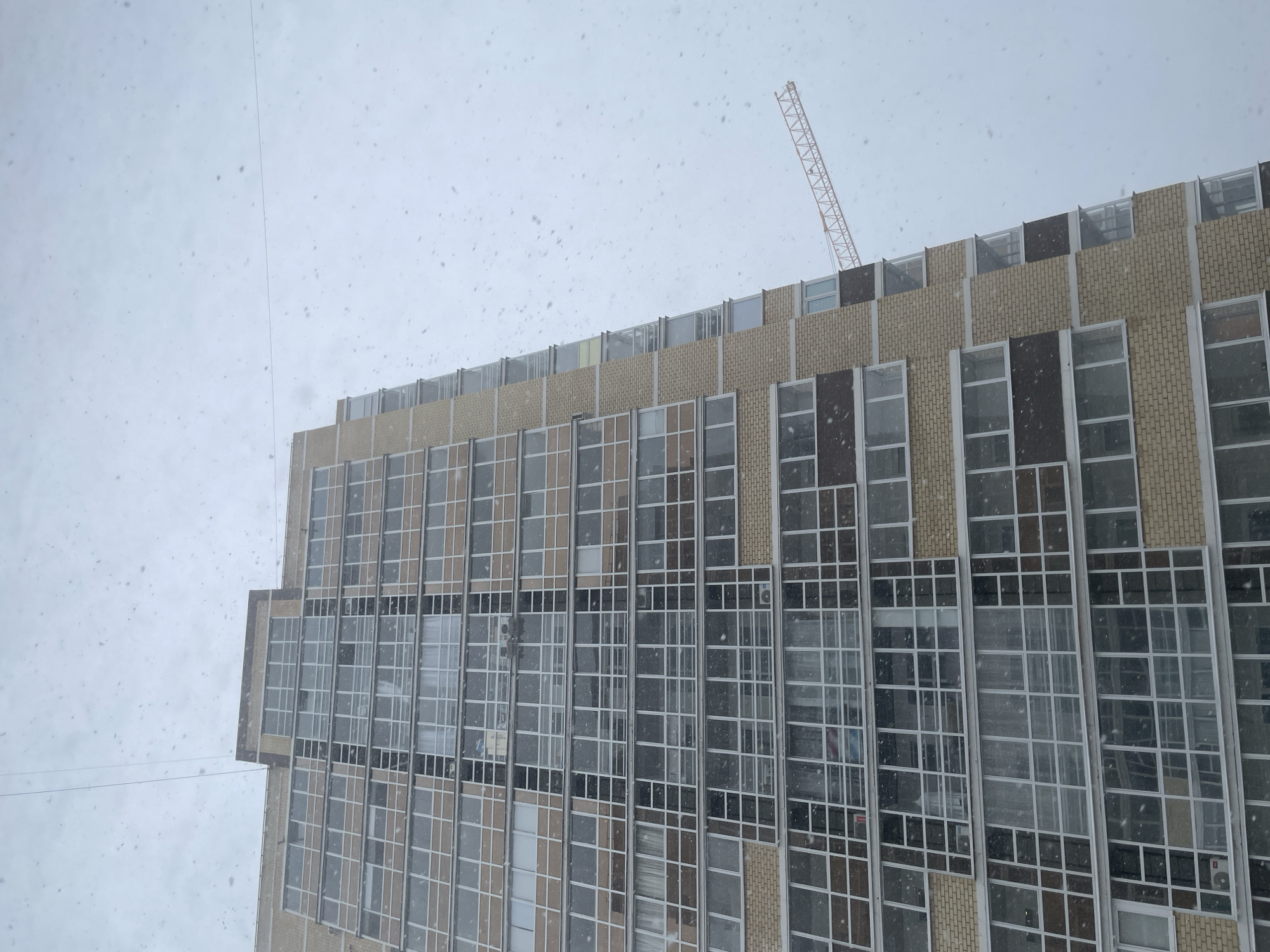 The image depicts a multi-story building, possibly located in Izhevsk, Russia, given the architectural style. The building's exterior consists of a repeating pattern of windows and brick or stone accents. A construction crane is visible atop the building. The weather appears snowy, with snowflakes visible in the foreground and background. No people are visible in the image.