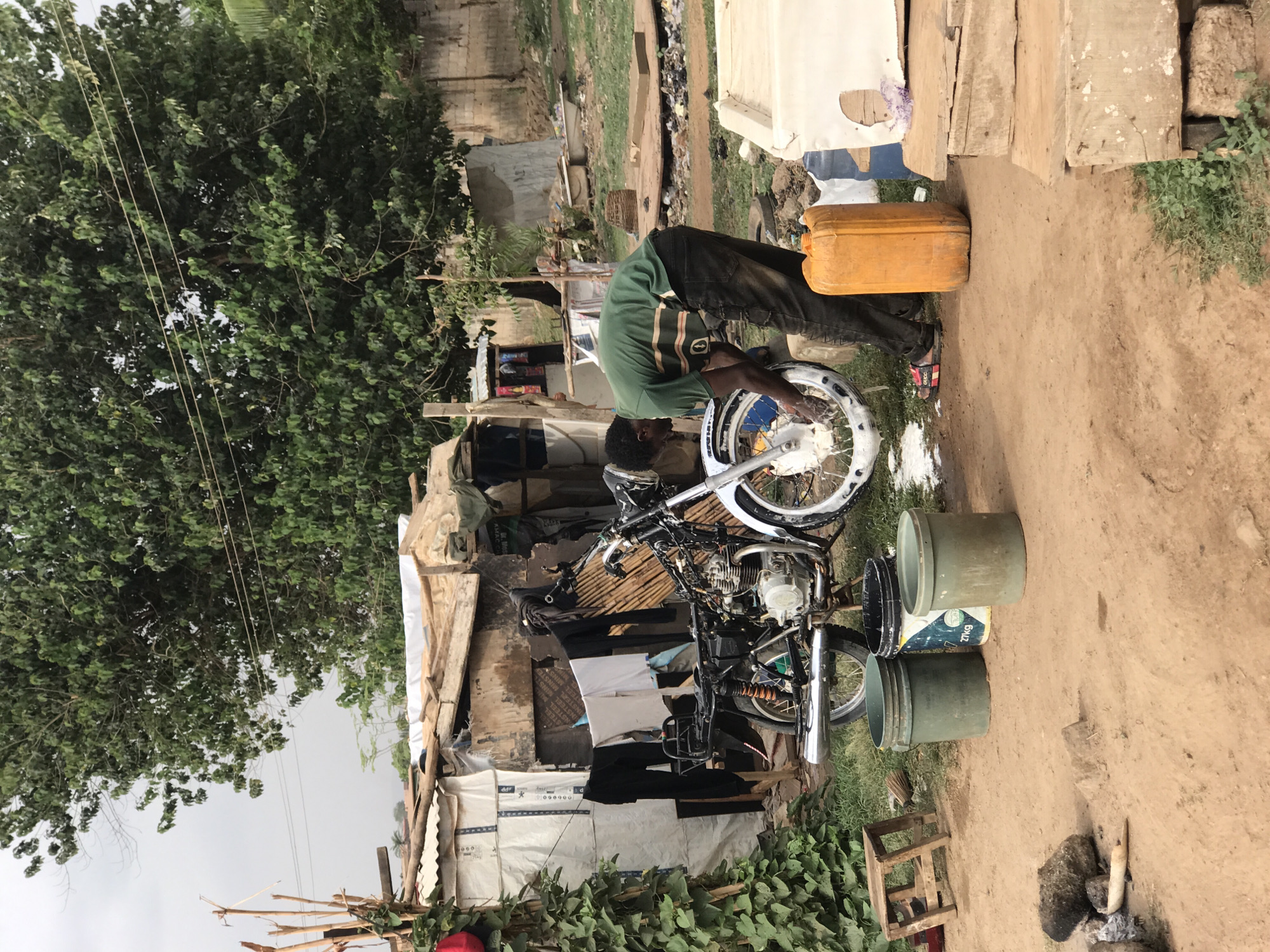 A man is washing the front wheel of a motorcycle in what appears to be an informal, possibly impoverished setting. He is wearing a green shirt with brown stripes, dark pants, and sandals. He is bent over, working on the wheel with a cloth or sponge. A yellow container and several buckets are nearby, likely containing water and cleaning supplies. The motorcycle is black and silver. Behind the man and the motorcycle, there are makeshift structures constructed from various materials including wood, tarps, and metal sheets. Clothes are hanging to dry. The ground is dirt and appears to be outdoor. There is a large tree to the left and an unkempt area with trash and debris in the background. It is not possible to confirm the location in the city of Abuja, Nigeria from the provided image.