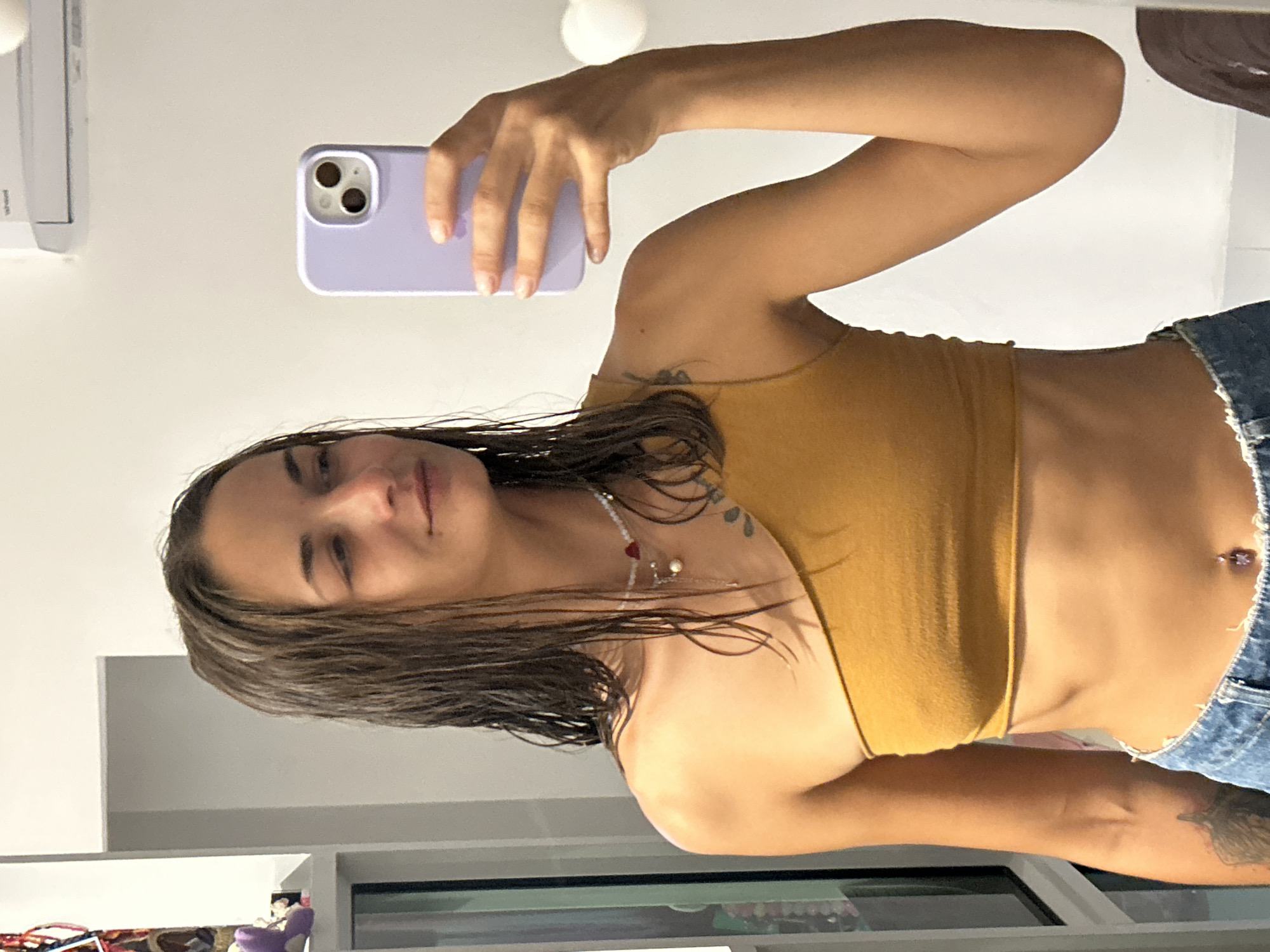 A woman is taking a mirror selfie in Kathu, Thailand. She has wet, brown hair, a bare midriff, and is wearing a mustard-yellow one-shoulder top and denim jeans. She is holding a light purple phone in her right hand. The background is a plain white wall with some shelving and a window visible on the left.