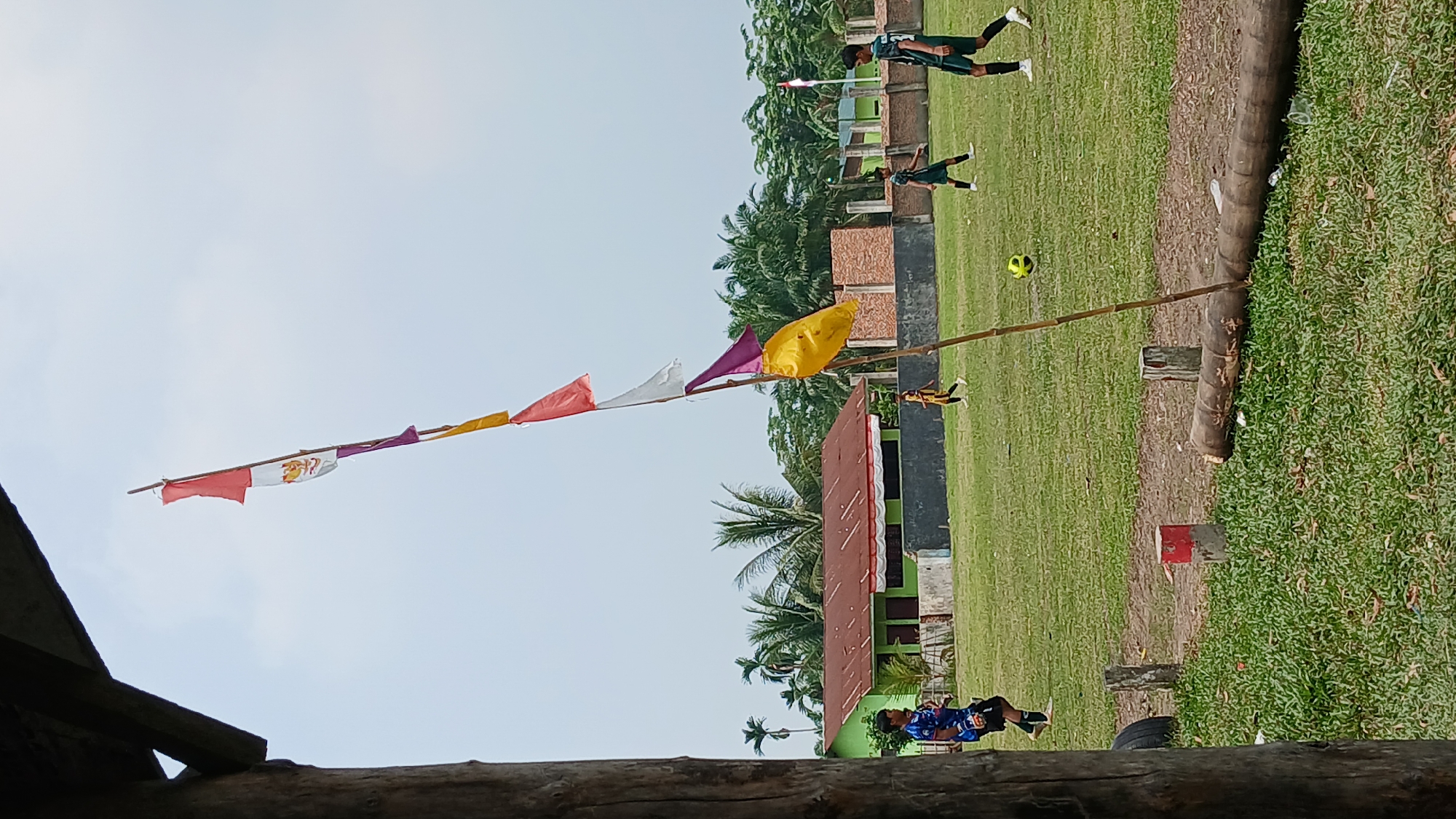 In Unknown, Indonesia, a soccer game is taking place on a grassy field. Three young men in soccer uniforms are visible on the field. One is wearing a blue and black jersey with white trim, black shorts, and white socks. Another is in a dark green jersey with white numbers, dark green shorts, and white socks. The third is wearing a dark green jersey with white numbers, dark green shorts, and white socks. A yellow soccer ball is on the field between two players. In the background, a green building with a red roof is visible, along with palm trees and other vegetation. A bamboo pole with several colored flags, including red, white, purple, and yellow, stands in the foreground. The sky is overcast.