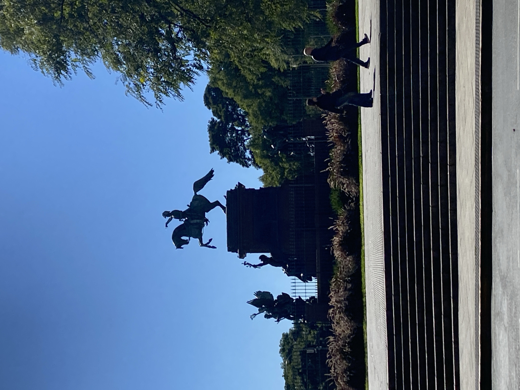 The image depicts a sunlit outdoor scene in Buenos Aires, Argentina, featuring a large public monument and people walking. The sky is a vibrant, clear blue, indicating fair weather during the daytime, likely morning or late afternoon given the distinct shadows.
On the left side of the frame, a grand bronze monument is silhouetted against the bright sky. The most prominent feature is a large equestrian statue of a figure on horseback, with other smaller statues and sculptural elements surrounding its base. These additional figures appear to be part of a larger allegorical or historical composition, characteristic of monuments found in public squares. Green trees and manicured bushes provide a natural backdrop, suggesting a park or a landscaped area.
In the foreground and extending towards the right, there is a wide paved area, possibly steps or a decorative pathway, characterized by strong parallel lines created by alternating light and shadow. Several people are visible walking along this path, also appearing somewhat silhouetted against the bright ground. Two individuals are clearly distinguishable in the upper right portion, walking away from the viewer.
The scene conveys a calm, everyday atmosphere, with visitors or passersby moving through a significant public space. No visible text is present in the image.